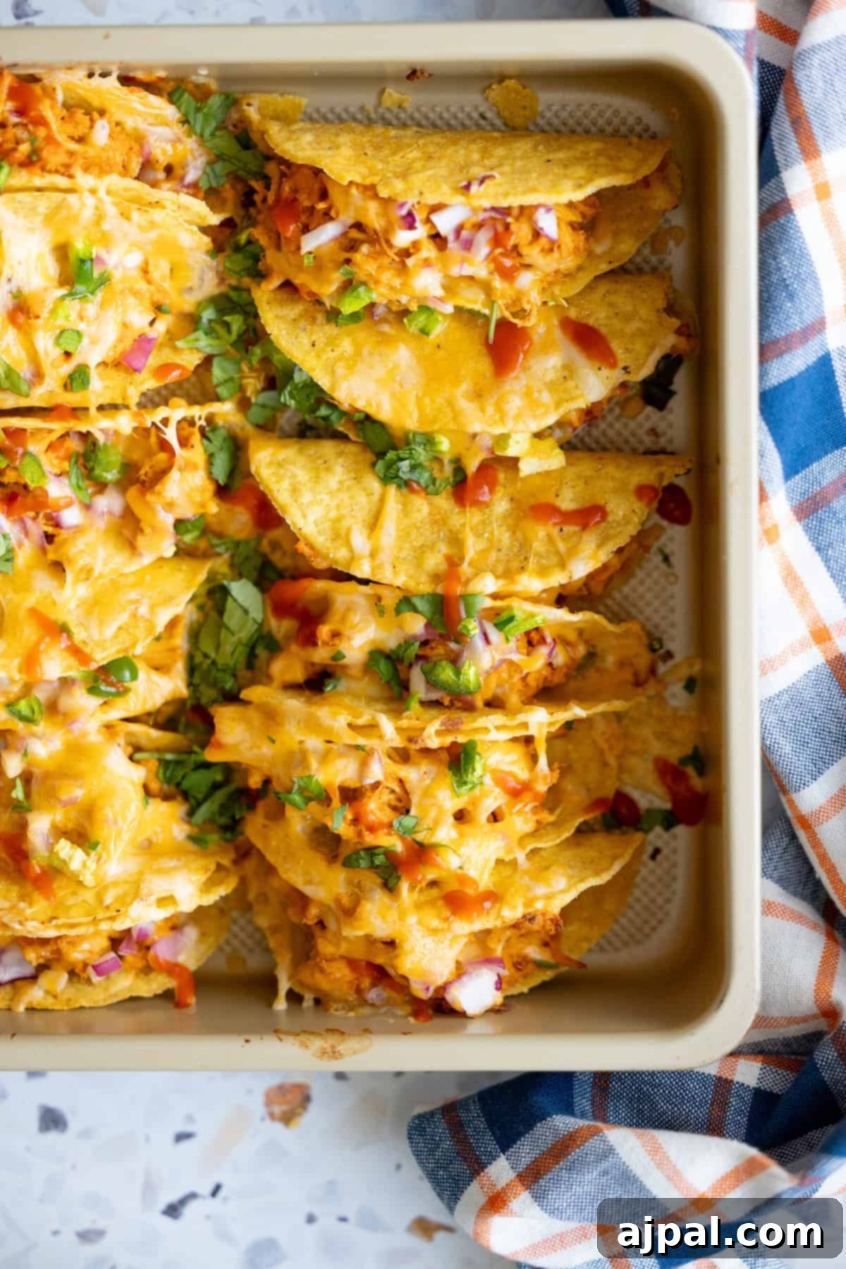 A baking tray filled with baked chicken tacos topped with melted cheese, diced red onions, chopped green herbs, and sauce, next to a blue and orange plaid cloth.