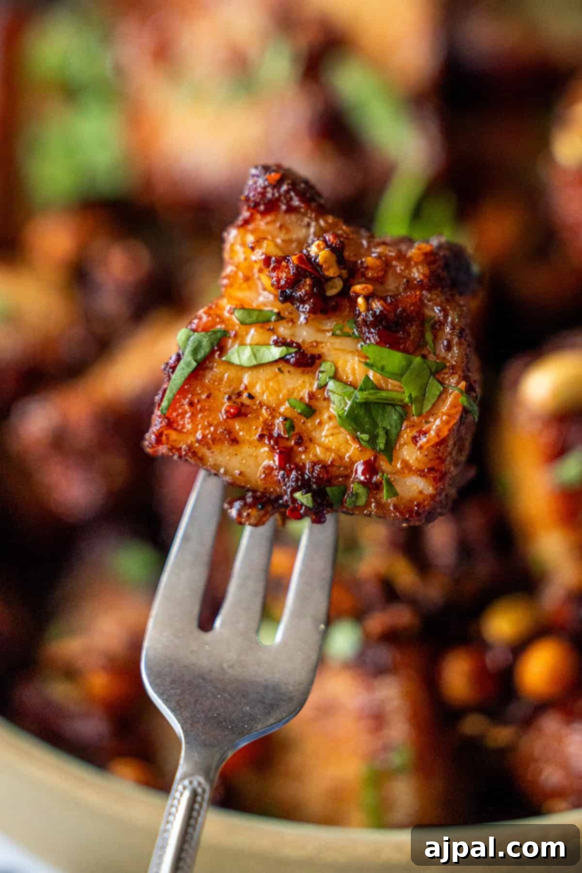Glazed Asian Pork Belly Morsels 3 A close-up of a fork holding a piece of Sticky Asian Pork Belly, roasted and seasoned to perfection, garnished with chopped herbs.