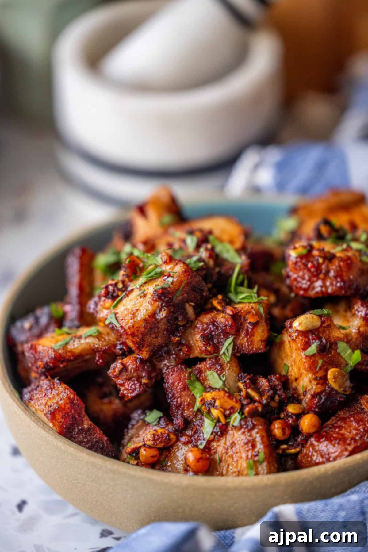 Glazed Asian Pork Belly Morsels 4 A bowl of crispy Sticky Asian Pork Belly pieces, seasoned and garnished with chopped herbs and spices, set on a marble surface next to a mortar and pestle.