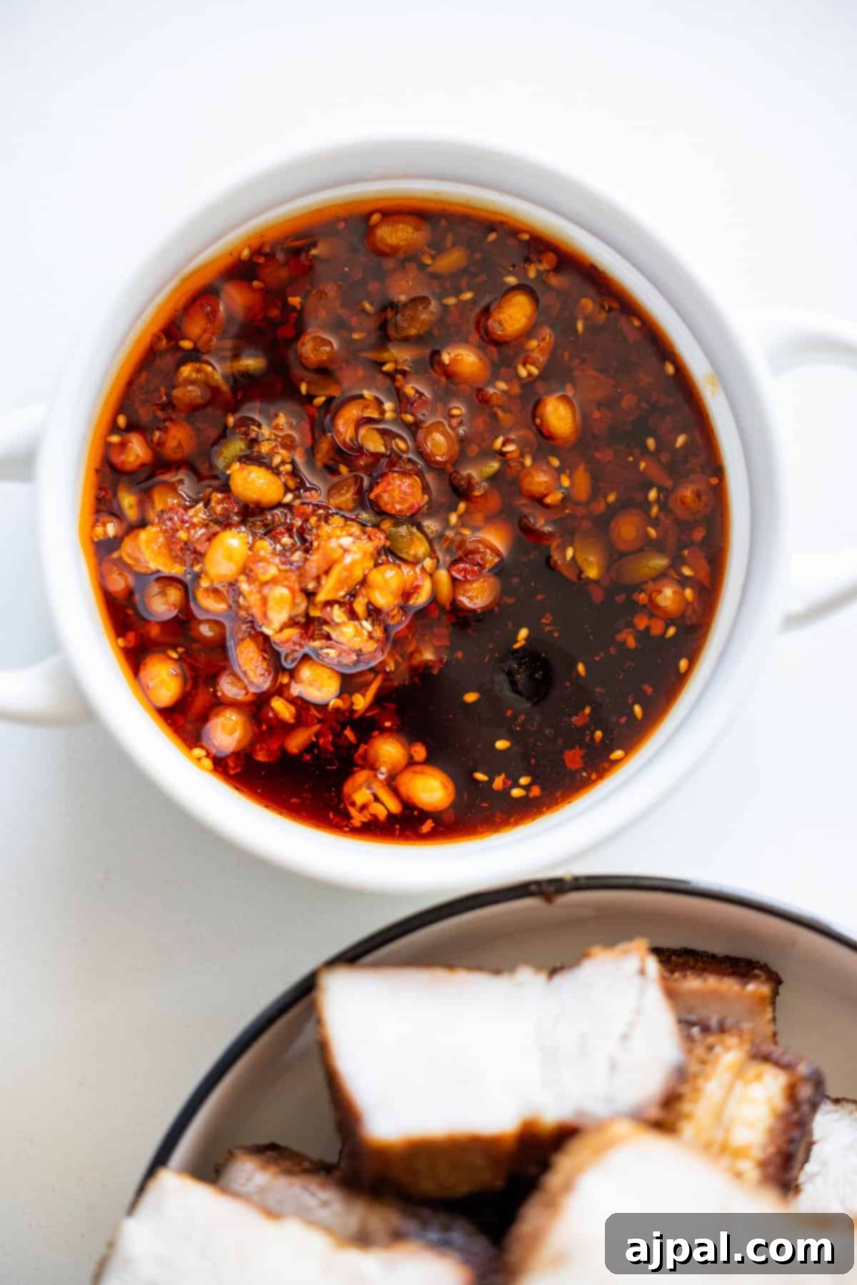 Glazed Asian Pork Belly Morsels 6 A white bowl filled with chili oil containing chili flakes and seeds sits next to a bowl of Sticky Asian Pork Belly, featuring perfectly sliced roasted pieces.