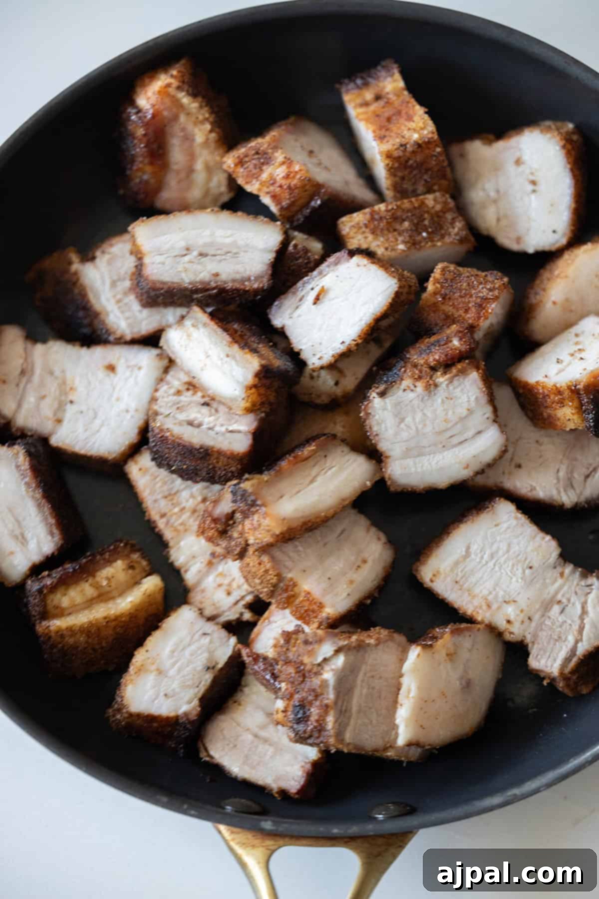 Glazed Asian Pork Belly Morsels 7 Sliced pieces of Sticky Pork Belly with crispy, browned edges are arranged in a black skillet with a gold handle.