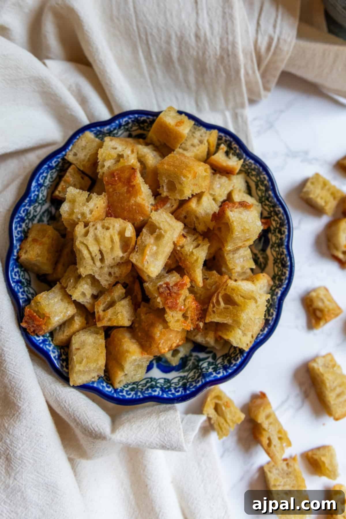 Crunchy Bread Bites 3 A decorative blue bowl filled with golden-brown homemade croutons sits on a light cloth, with a few croutons scattered nearby on a marble surface.