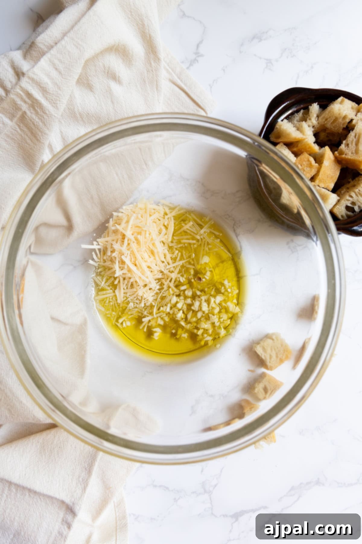Crunchy Bread Bites 5 A glass bowl contains olive oil, grated Parmesan cheese, and minced garlic on a marble surface, with a small dish of homemade croutons nearby.