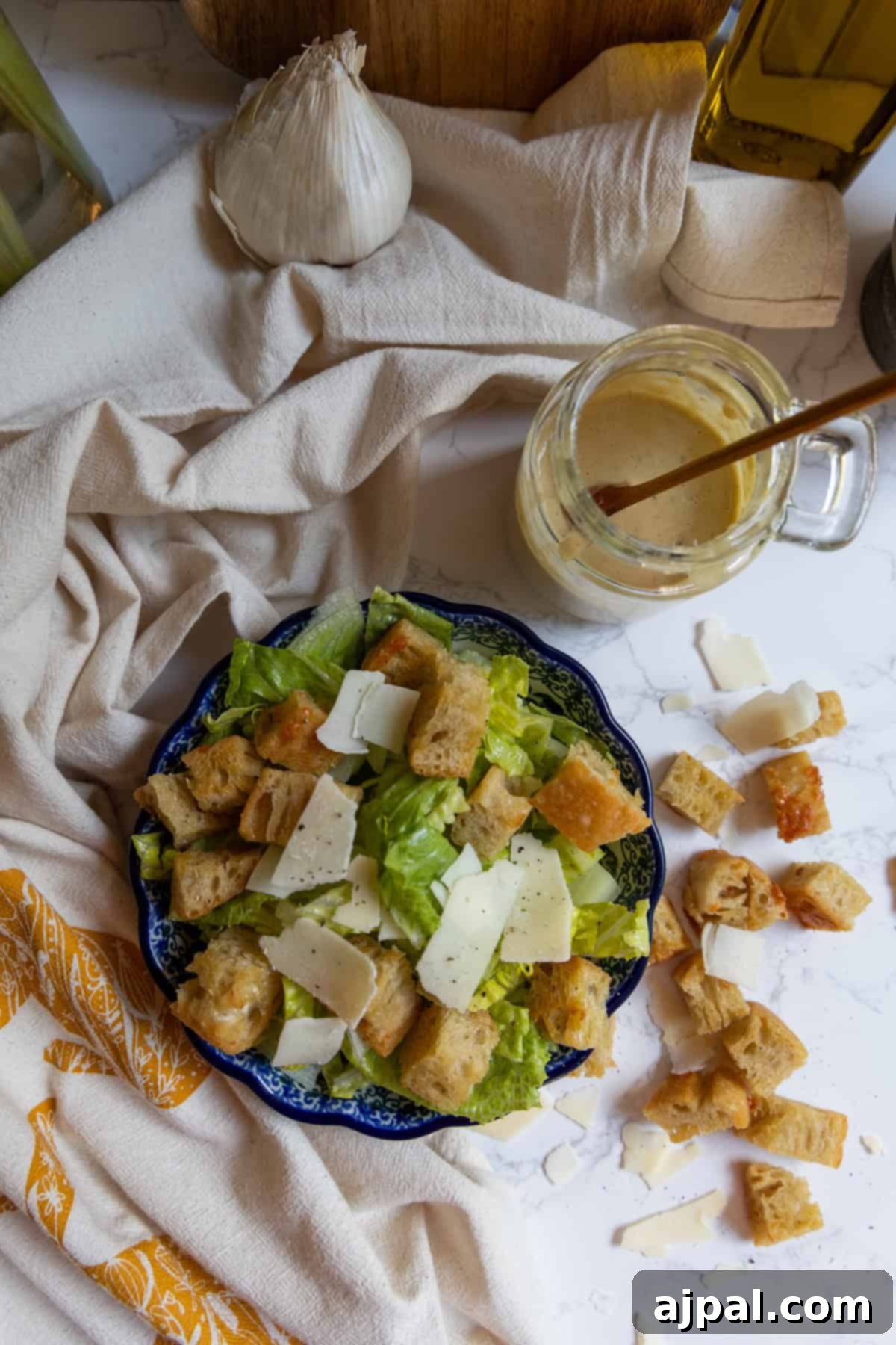 The Ultimate Caesar Salad 7 A vibrant bowl of Caesar salad, abundant with crisp romaine, golden croutons, and elegant shaved Parmesan, rests on a textured cloth. Accompanying it are a bulb of garlic, a jar of homemade Caesar dressing with a spoon, and scattered croutons and cheese, highlighting the fresh, prepared ingredients.
