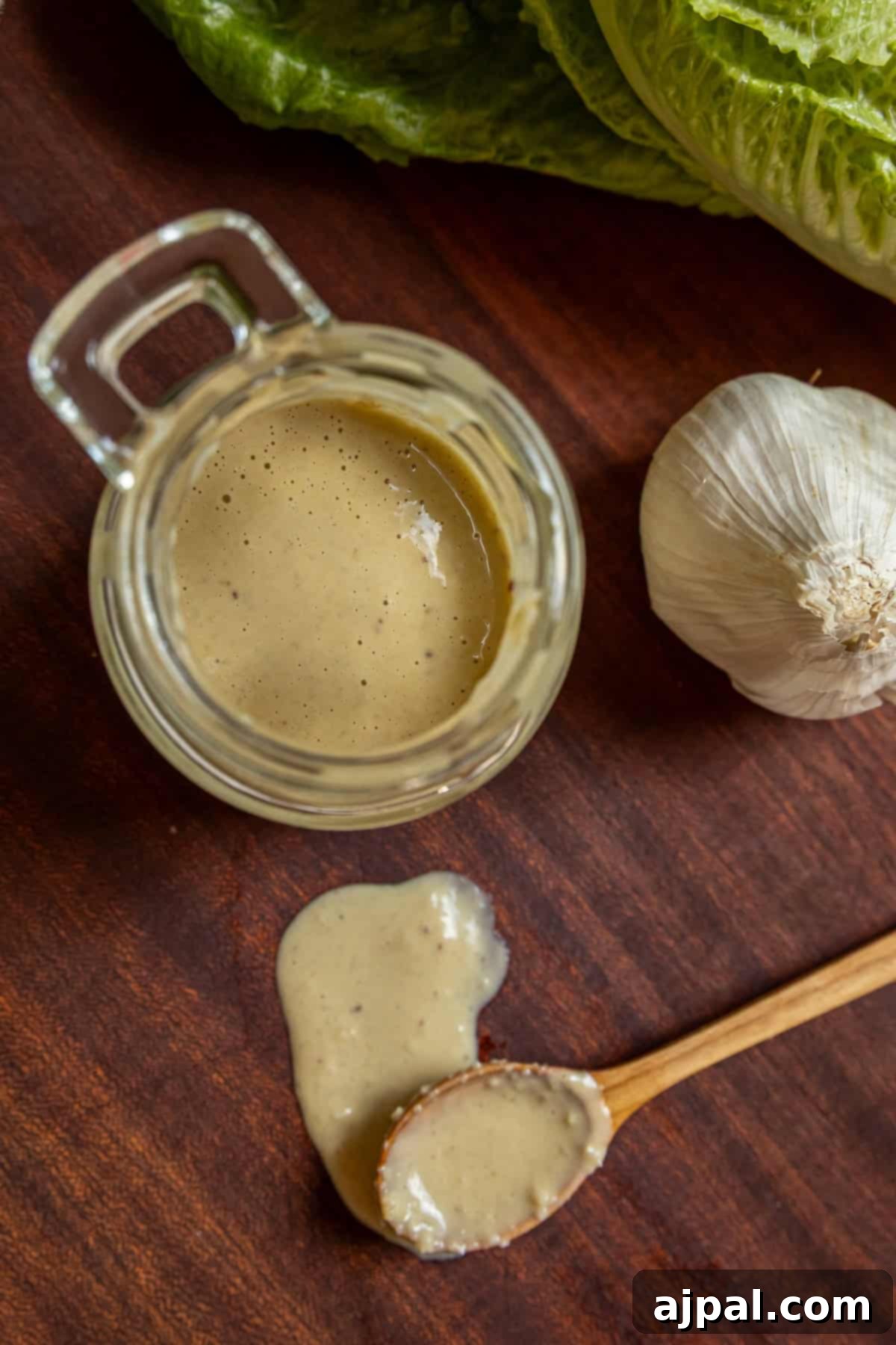 A jar of creamy homemade Caesar dressing sits on a rustic wooden table beside fresh garlic, crisp romaine lettuce, and a wooden spoon with spilled dressing, highlighting its fresh ingredients.