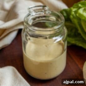 A glass jar filled with creamy caesar dressing sits on a wooden surface next to a lettuce leaf and a light-colored cloth.