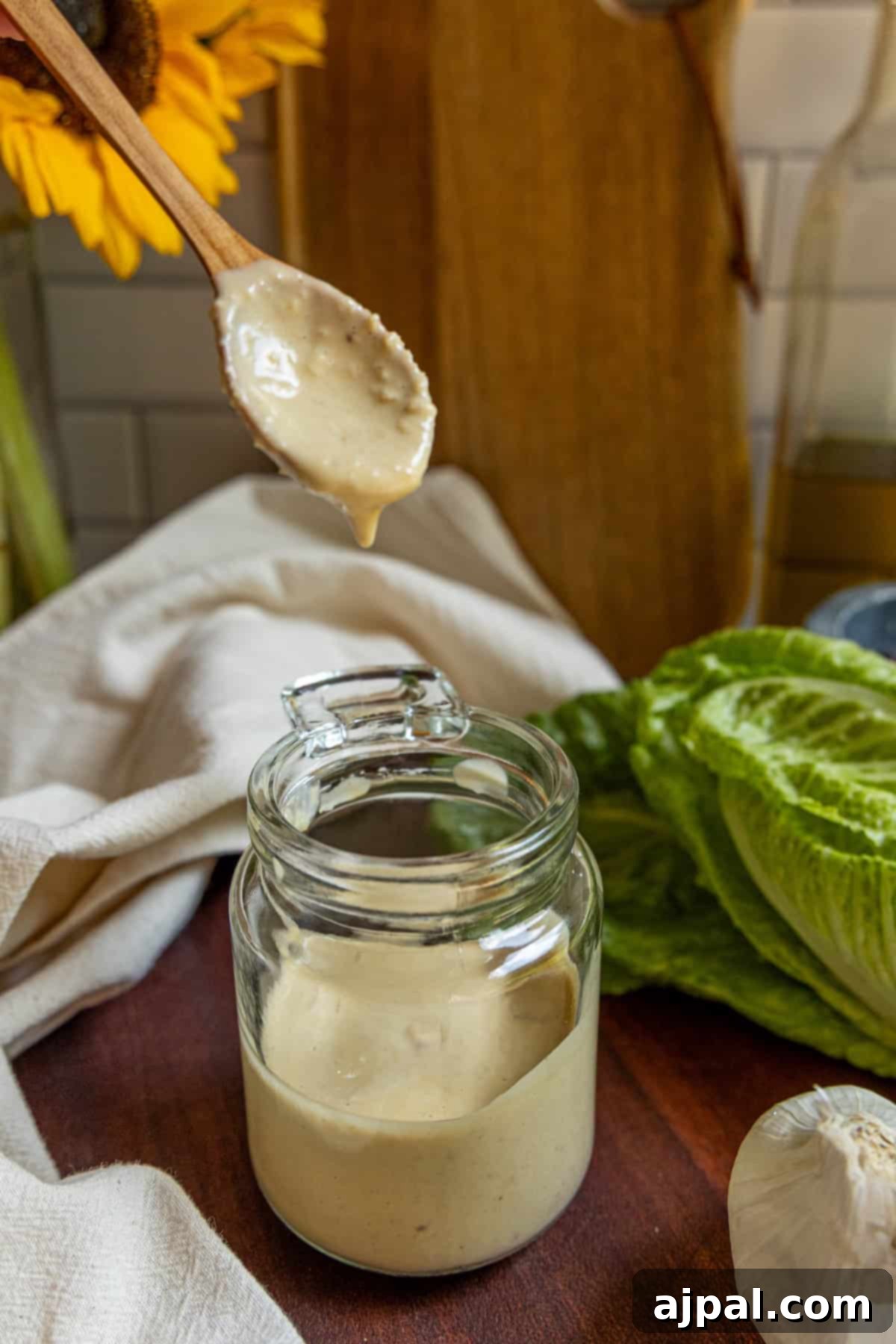 A wooden spoon with creamy caesar dressing hovers over an open glass jar; lettuce, garlic, and a cloth rest nearby on the wooden surface.