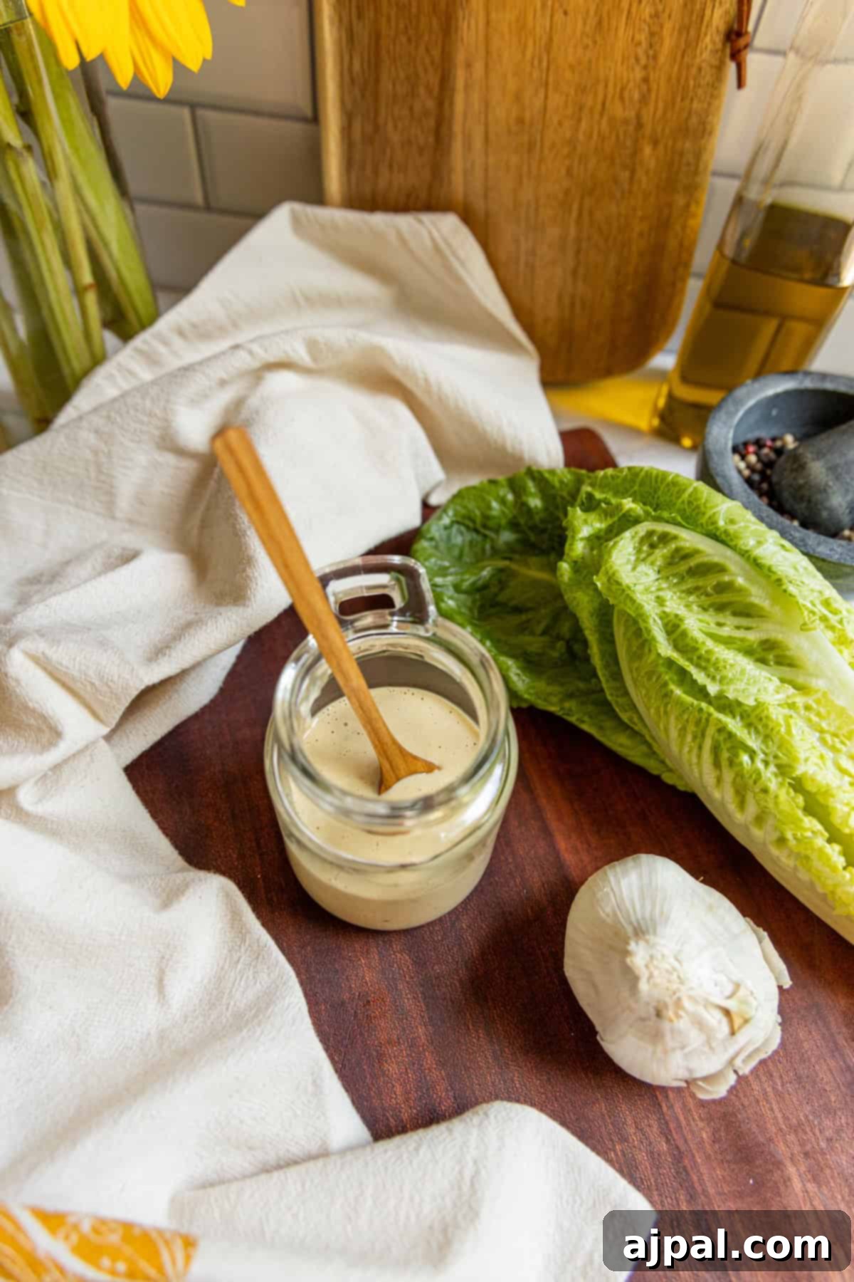 A jar of creamy homemade Caesar dressing with a wooden spoon, a head of fresh romaine lettuce, and a bulb of garlic rest on a rustic wooden surface, complemented by a white cloth, setting a natural and inviting scene.