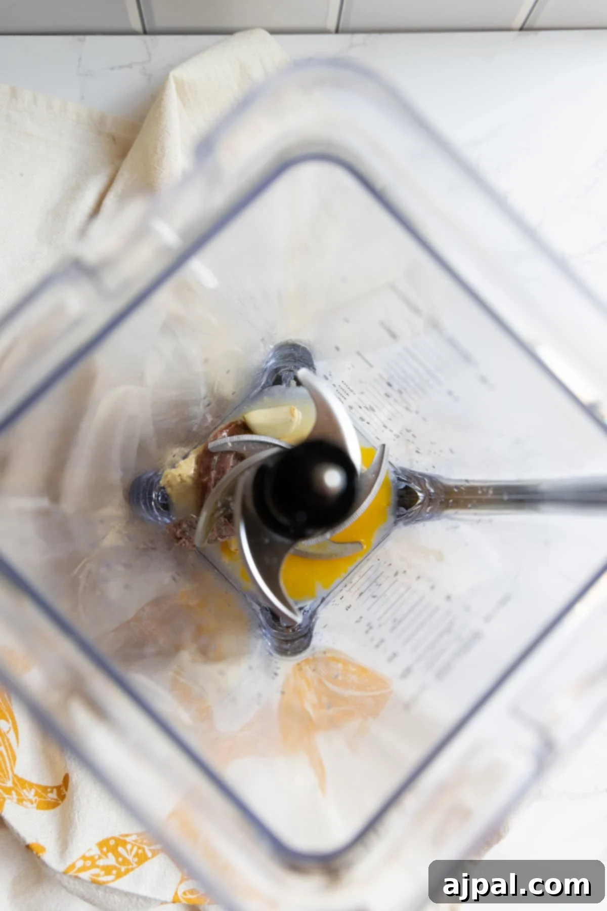 Top-down view of an empty blender, displaying faint streaks of creamy Caesar dressing residue on its inner sides. The blender sits on a clean white counter, partially resting on a soft beige and orange towel, suggesting recent use and easy cleanup.