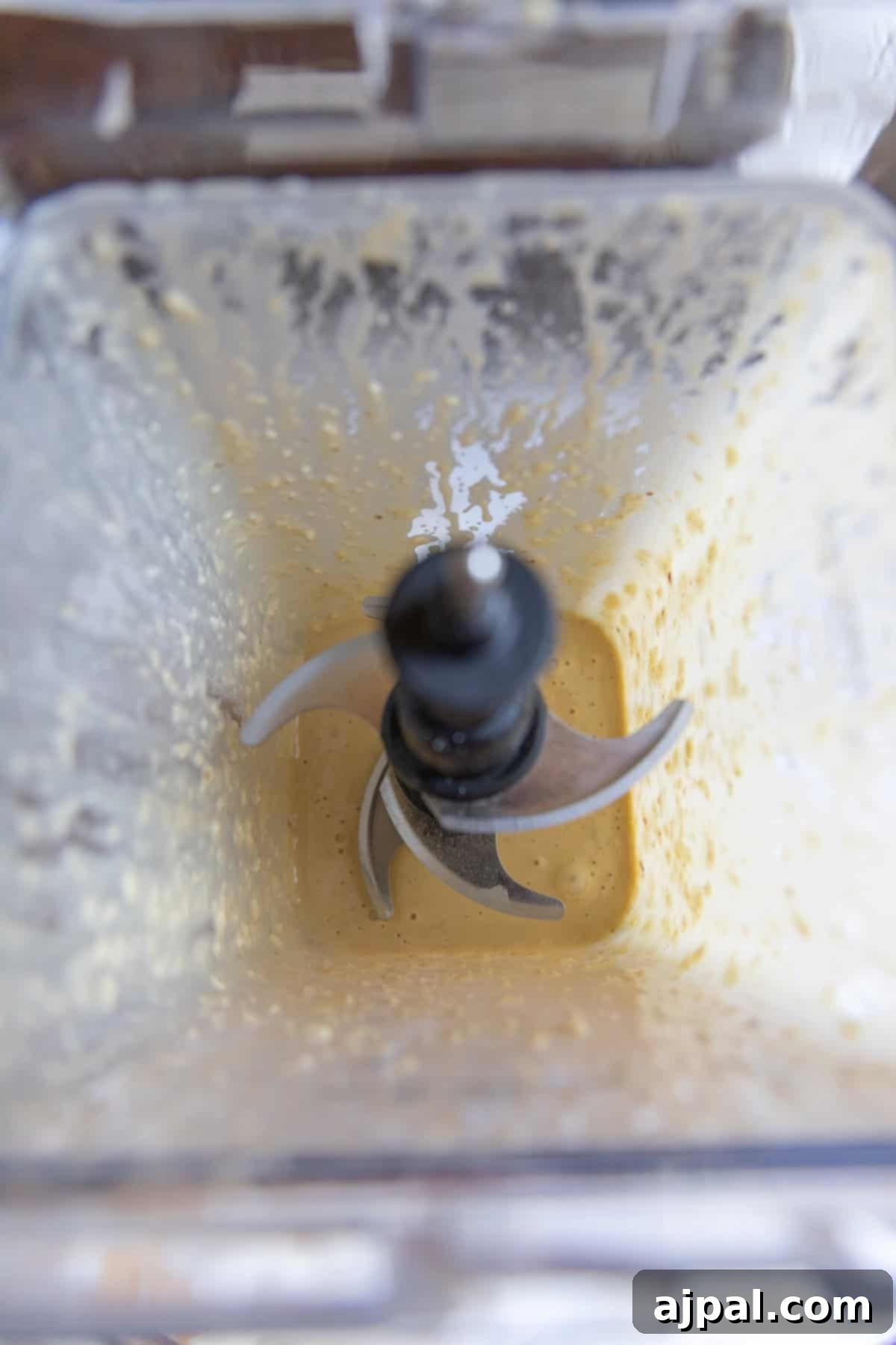 Top-down view of a blender containing a creamy, light yellow caesar dressing mixture with remnants stuck to the sides and the metal blades visible in the center.