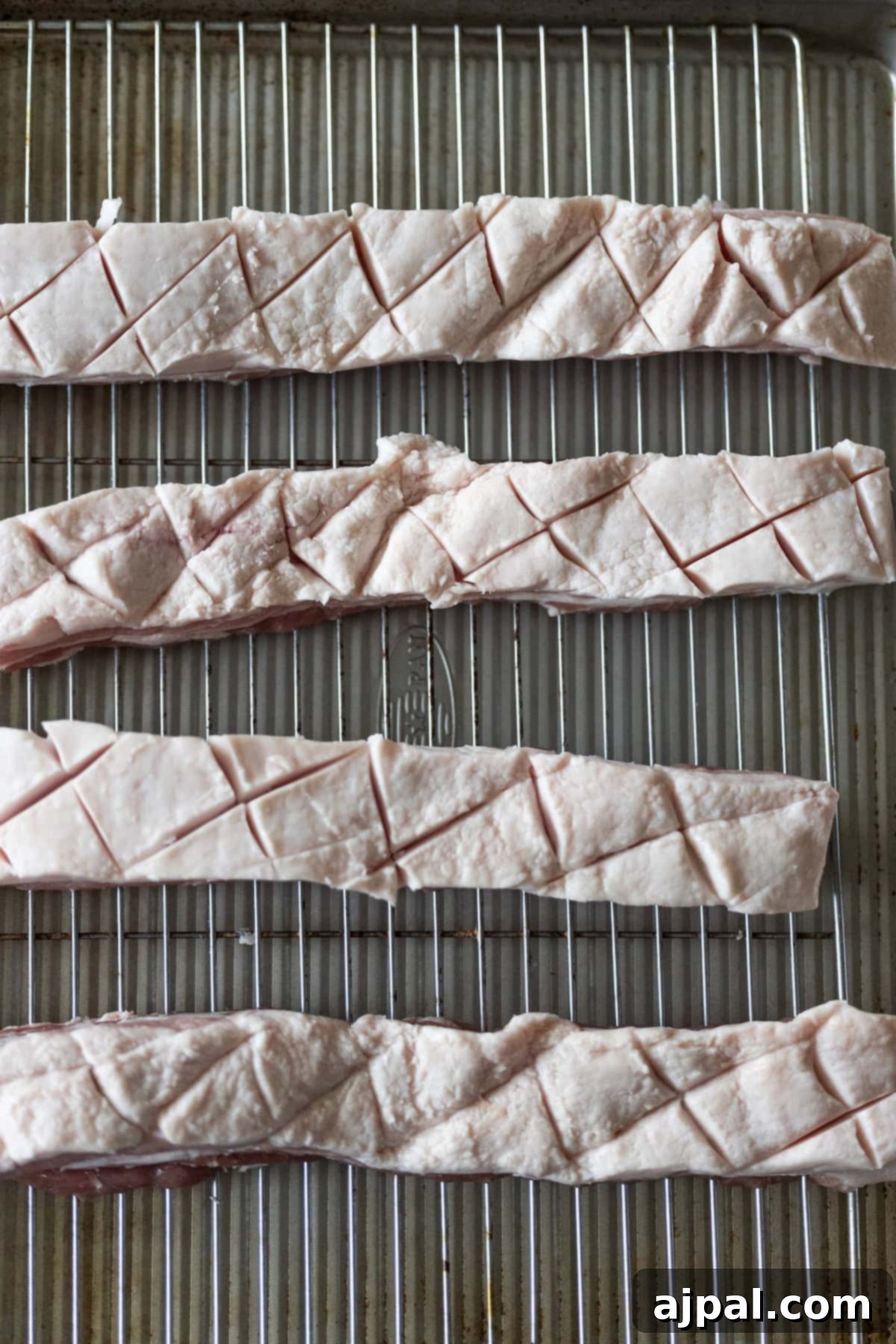 Ultimate Crispy Pork Belly 6 Four strips of raw roasted pork belly fat with a crosshatch pattern scored into the surface are arranged on a wire rack over a baking sheet.