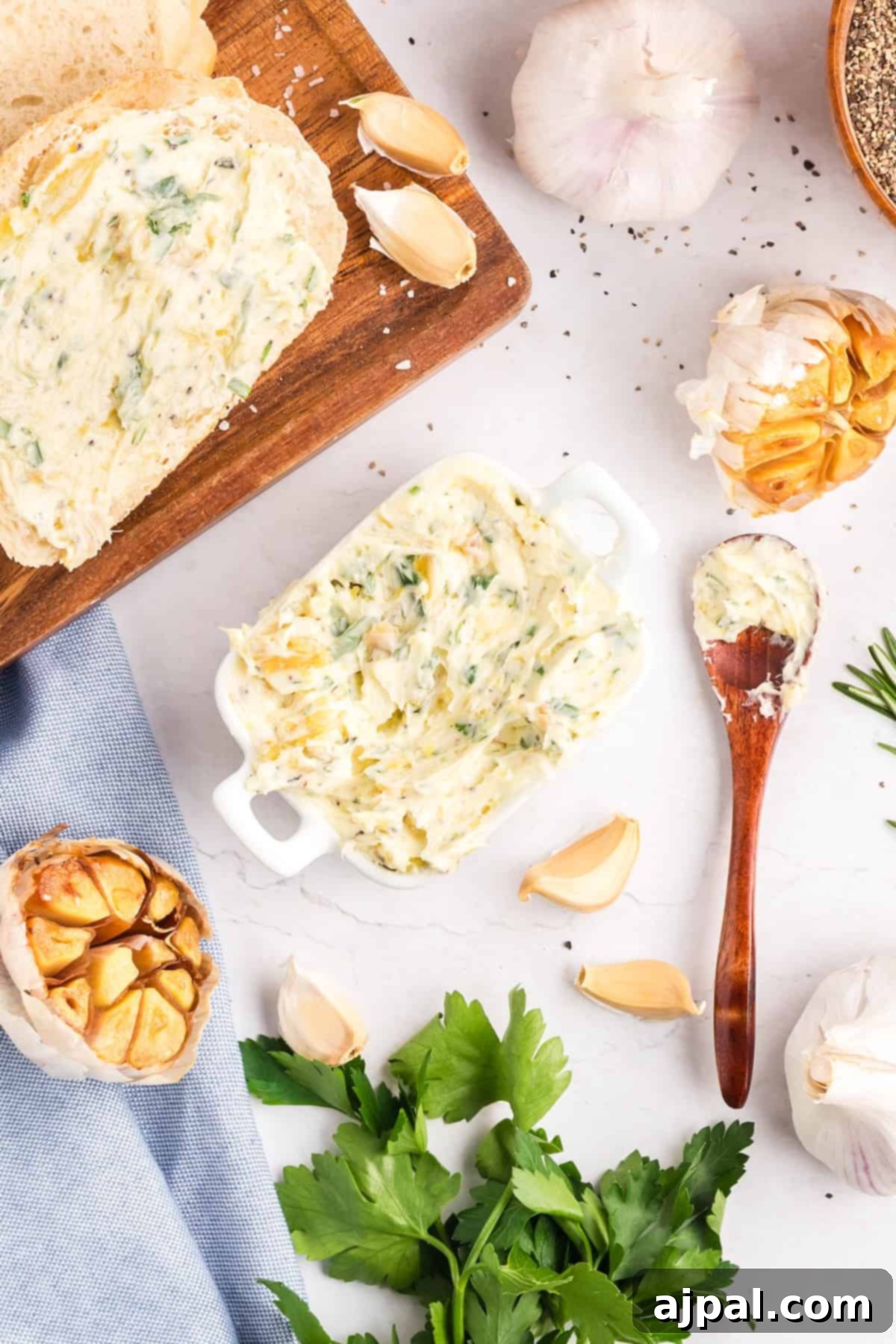 A small dish of roasted garlic butter with a wooden spoon, surrounded by parsley, garlic cloves, pepper, and sliced bread on a wooden board.