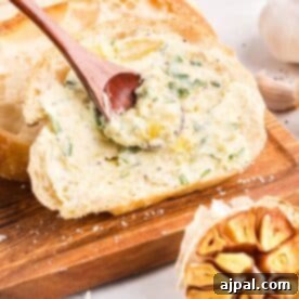 A slice of bread on a wooden board is being spread with rich roasted garlic butter using a wooden spoon, with a head of roasted garlic nearby.