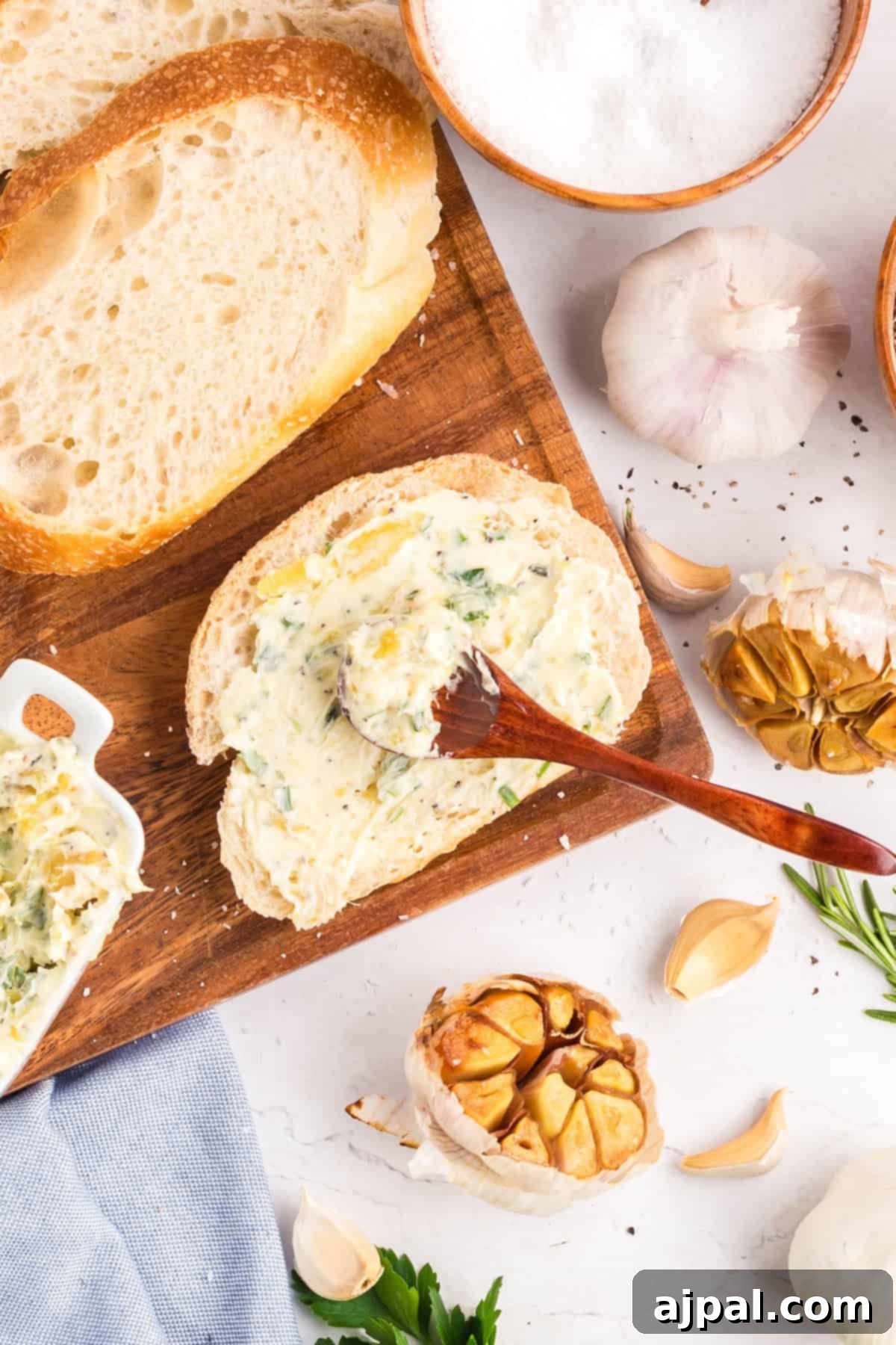 A slice of bread is spread with roasted garlic butter using a wooden spoon, surrounded by garlic, parsley, salt, and more bread on a wooden board.