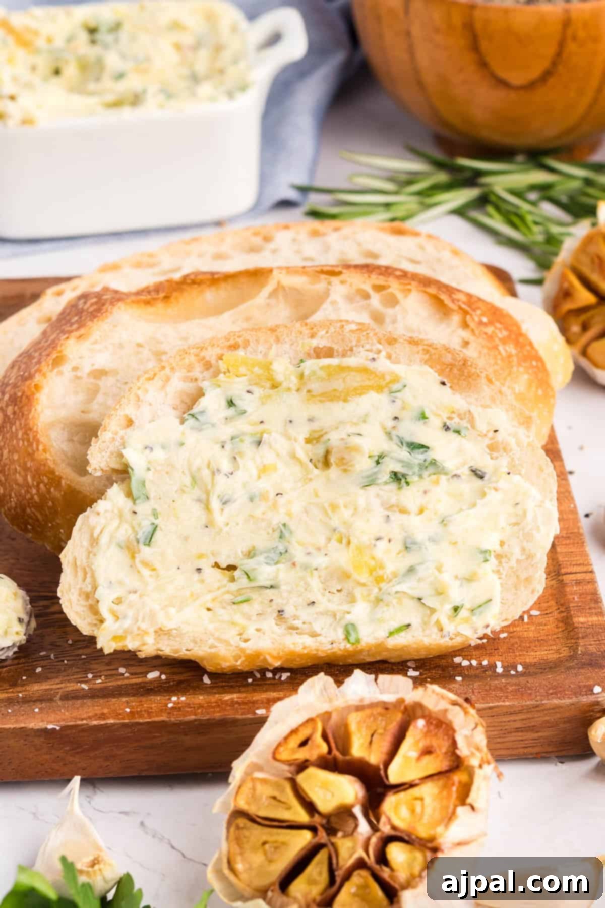 A slice of bread topped with roasted garlic butter rests on a wooden board, with a loaf of bread and roasted garlic nearby.