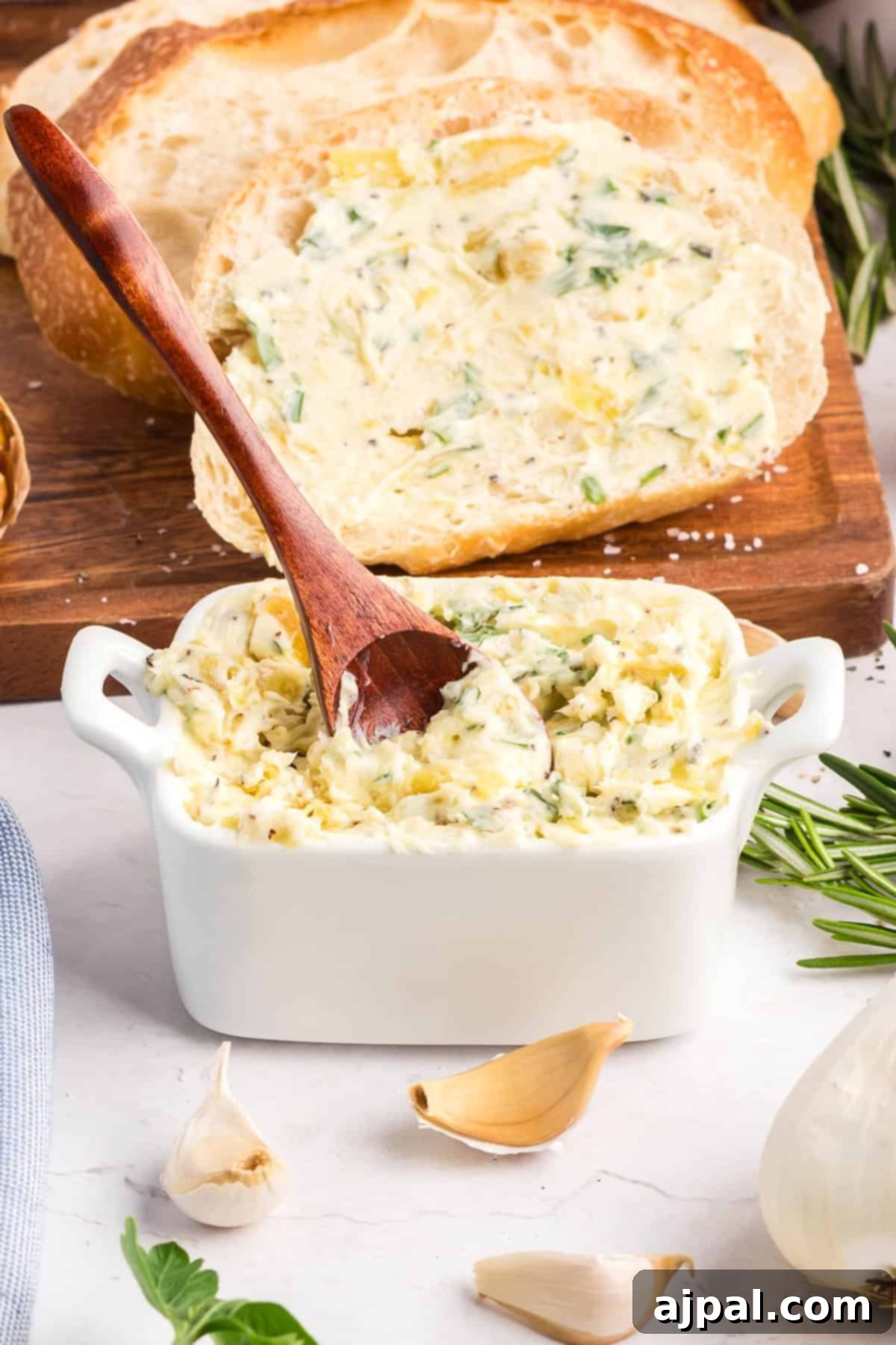A white dish filled with roasted garlic butter sits next to a wooden spoon, garlic cloves, and a slice of bread spread with the flavorful butter.