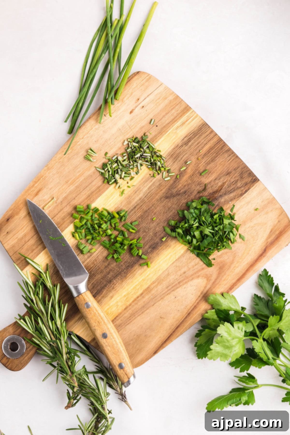 A wooden cutting board with chopped rosemary, chives, parsley, and hints of roasted garlic butter, alongside a knife, whole herbs, and green onions arranged on a white surface.