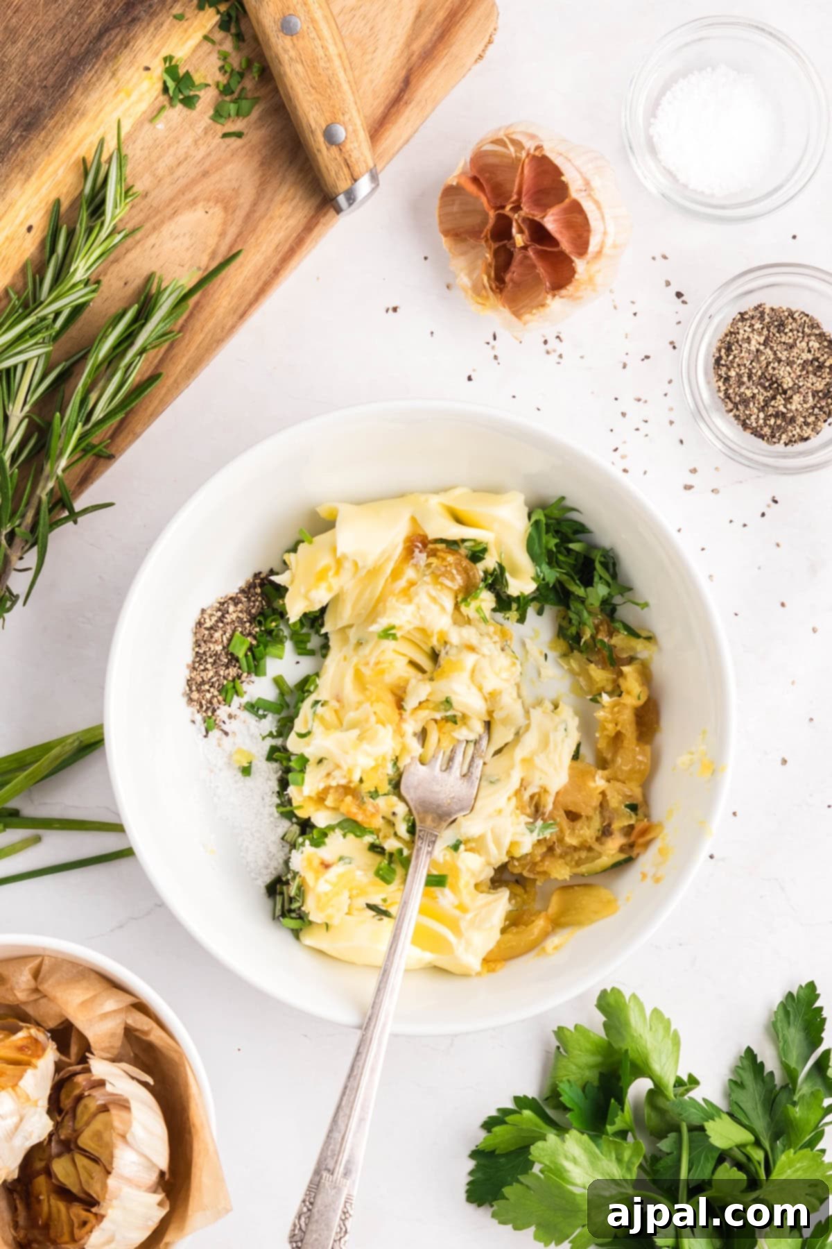 A bowl with softened butter, chopped herbs, roasted garlic, salt, and pepper is being mixed with a fork to create creamy Roasted Garlic Butter; fresh herbs and seasonings are arranged nearby on a white surface.