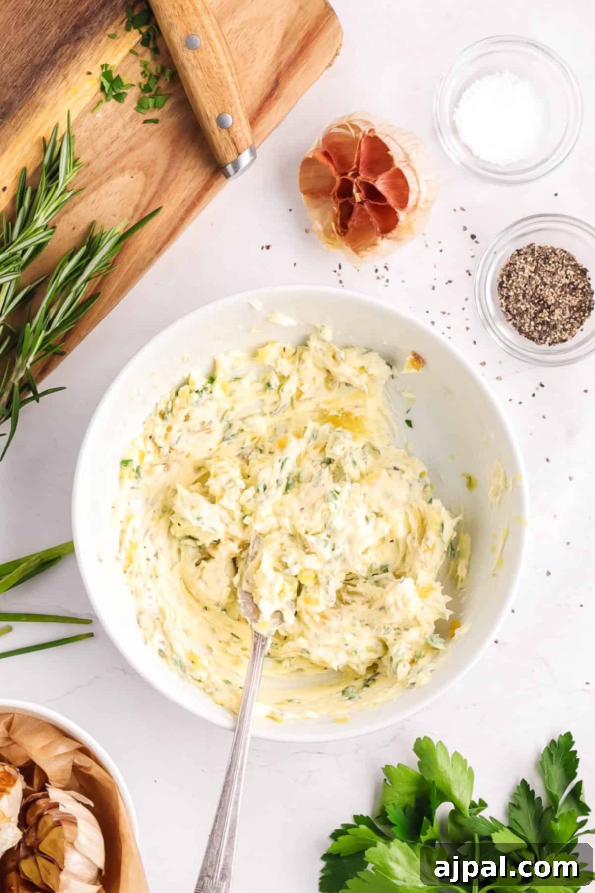 A bowl of roasted garlic butter with a spoon, surrounded by fresh herbs, garlic, salt, and pepper on a white countertop.