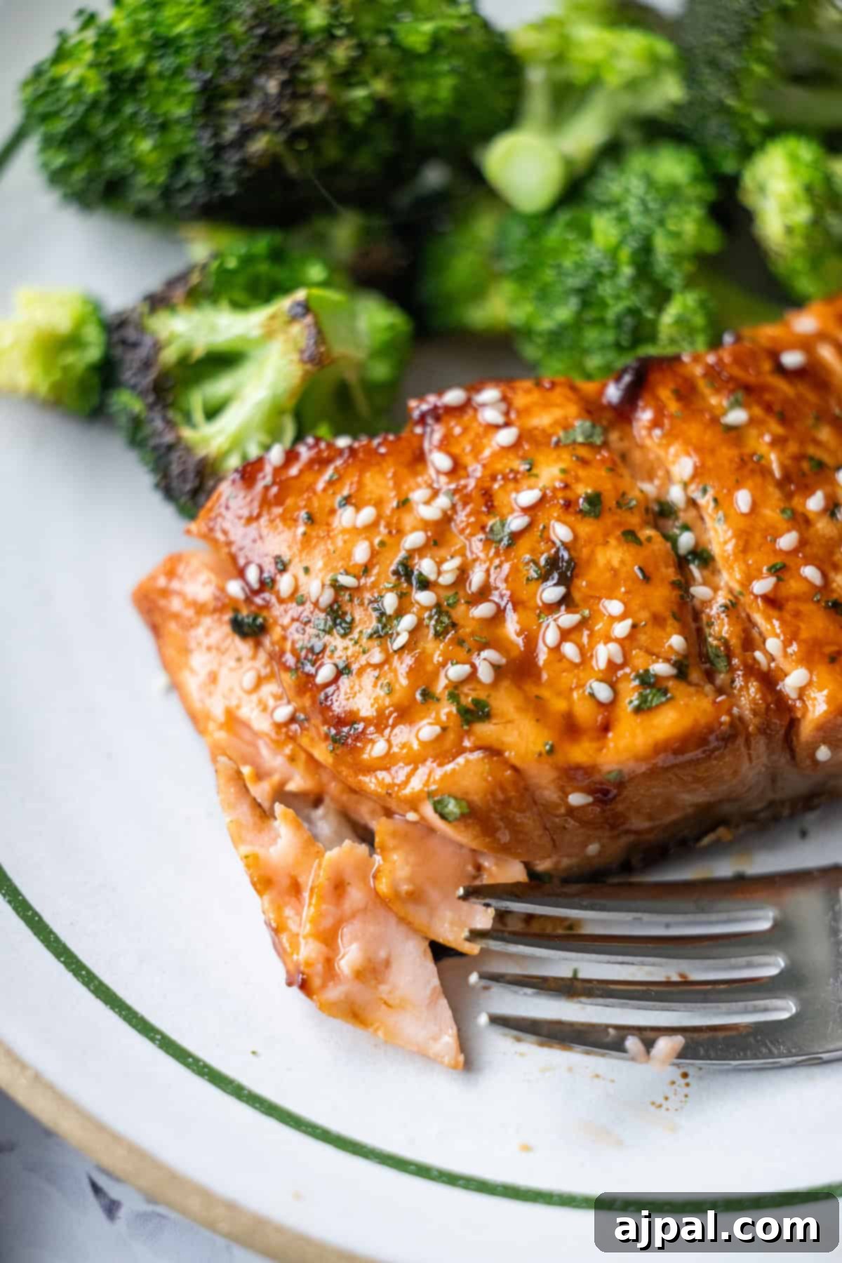 A close-up of Japanese BBQ Salmon, glazed and topped with sesame seeds and herbs, served with steamed broccoli on a white plate. A fork rests beside the salmon.