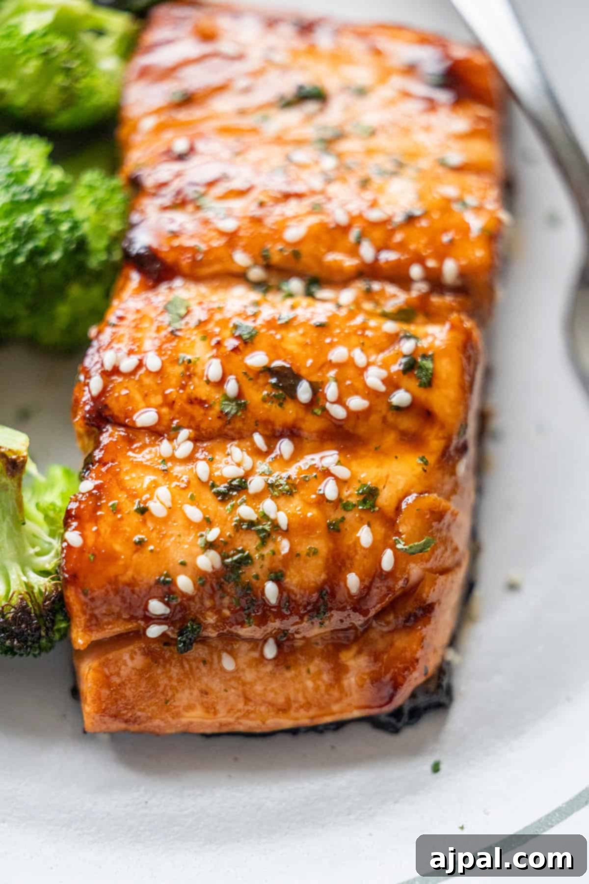 Close-up of Japanese BBQ Salmon fillets glazed to perfection, topped with sesame seeds and herbs, served with roasted broccoli on a white plate.