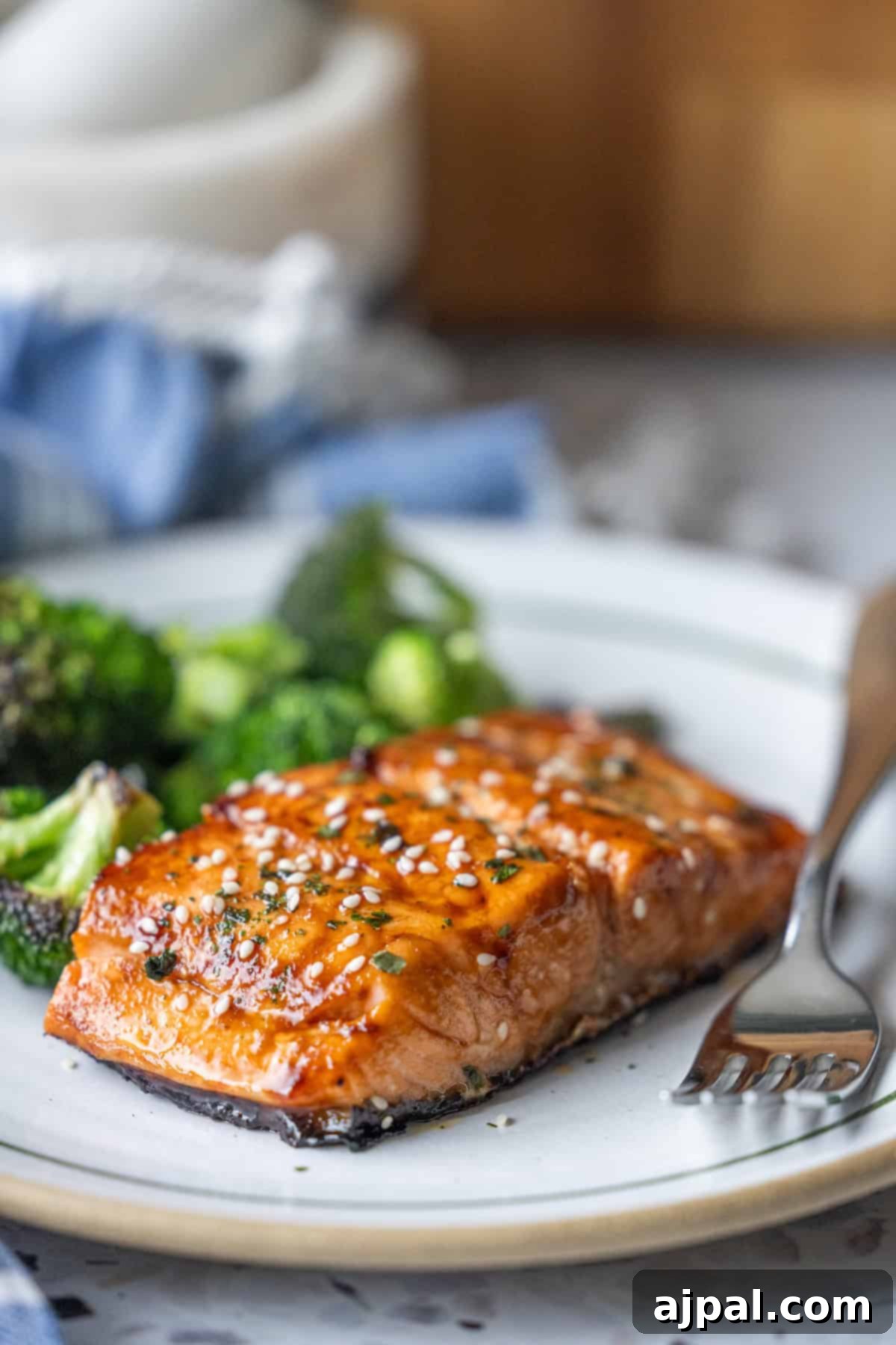 A Japanese BBQ Salmon fillet, garnished with sesame seeds and herbs, is served on a plate with a side of broccoli and a fork.