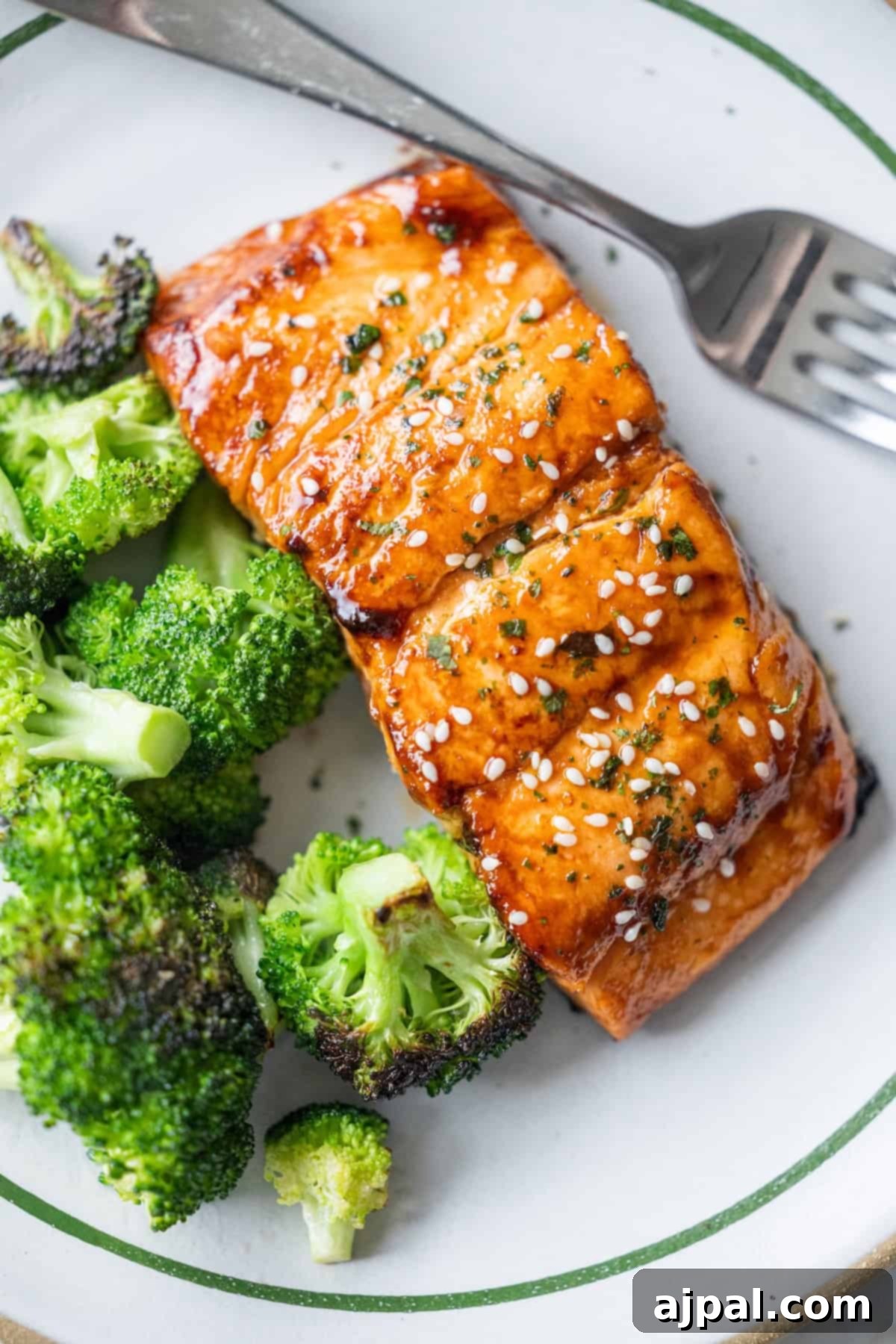 A plate with Japanese BBQ Salmon fillet glazed and topped with sesame seeds and herbs, served alongside roasted broccoli, with a fork on the side.
