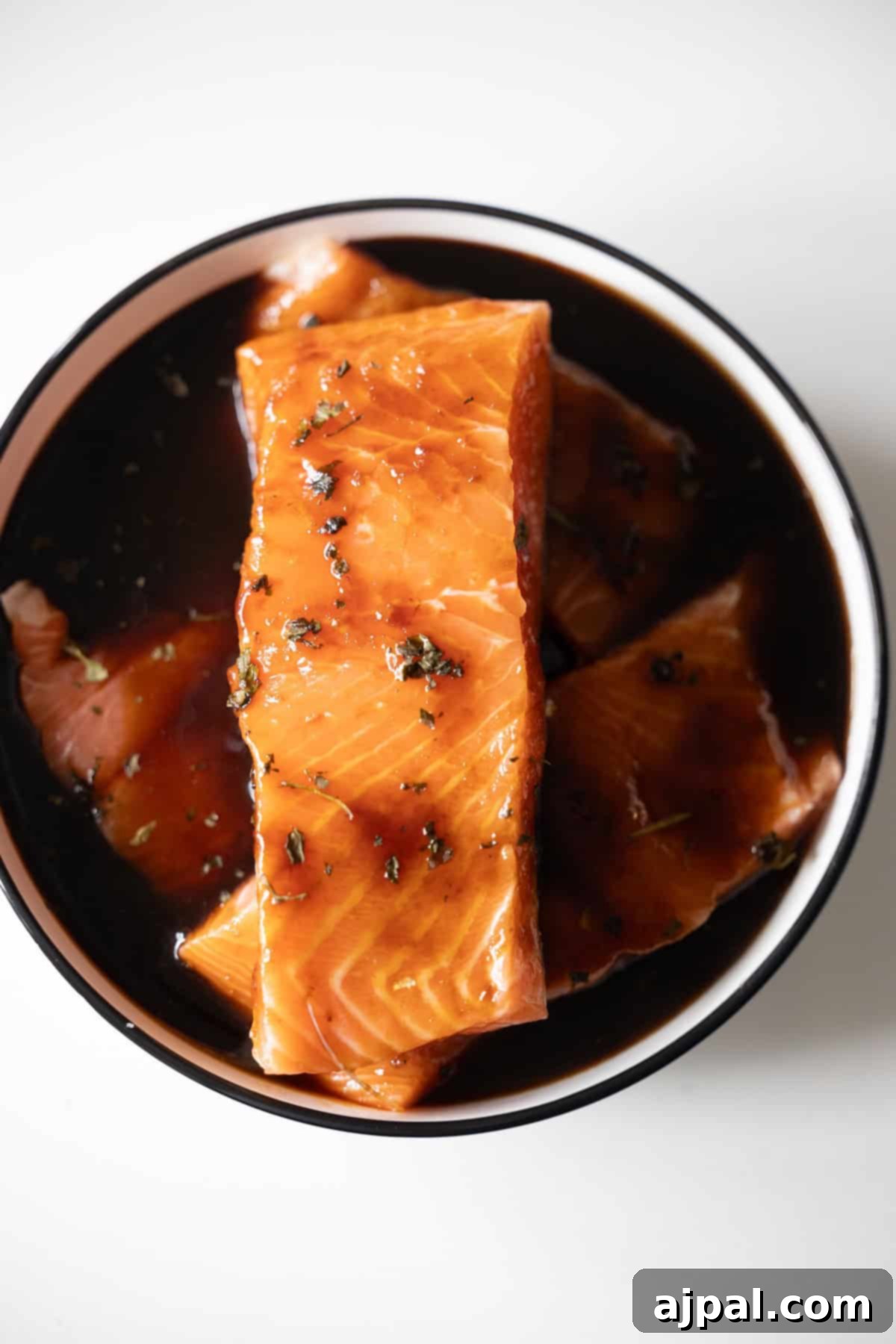 Japanese BBQ Salmon fillets marinate in a dark, flavorful sauce with herbs, arranged in a round bowl on a white surface.