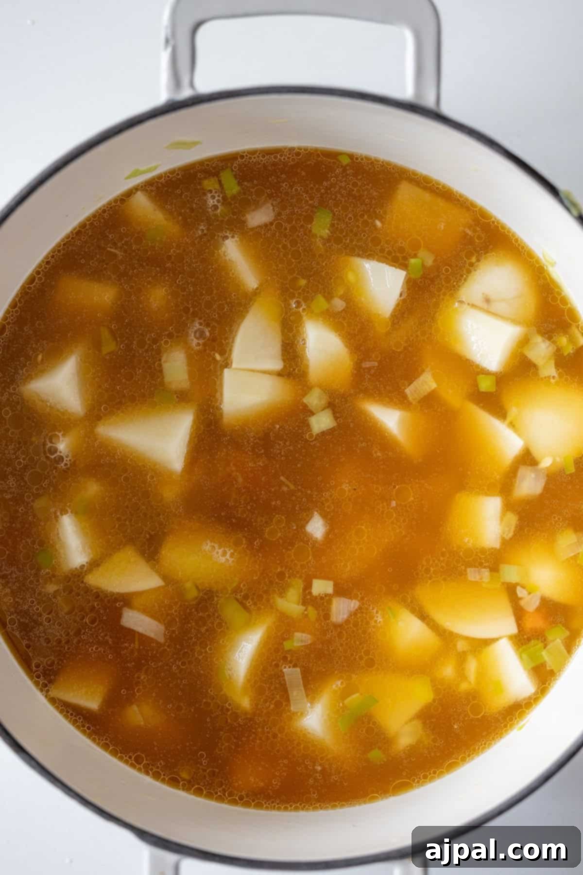 Traditional Finnish Salmon Soup 9 A pot filled with broth, chopped potatoes, onions, and celery on a white surface—ingredients ready to make Finnish Salmon Chowder, also known as lohikeitto.