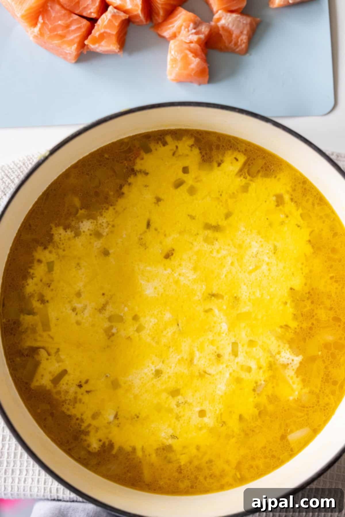Traditional Finnish Salmon Soup 10 A pot of yellow broth with chopped onions simmers on a stovetop, while diced raw salmon waits nearby—ingredients for a comforting Finnish Salmon Chowder, also known as lohikeitto.