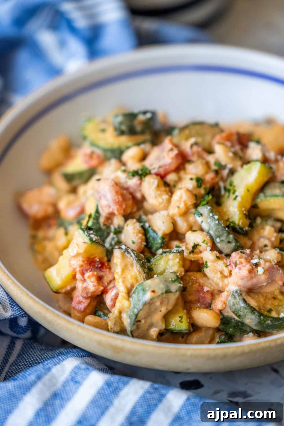 Marry Me Zucchini and Bean Delight 2 A bowl of creamy white beans mixed with Marry Me Zucchini, diced tomatoes, and fresh herbs, served on a blue and white striped cloth.