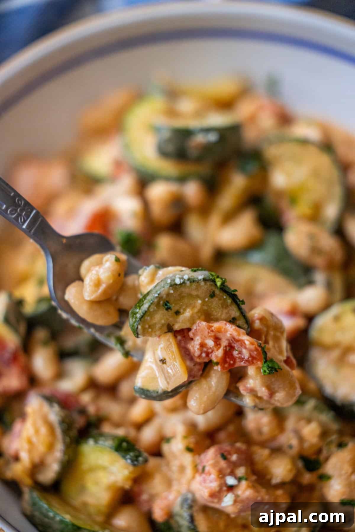 Marry Me Zucchini and Bean Delight 3 A close-up of a fork holding a bite of pasta with zucchini, white beans, tomatoes, and creamy sauce, with a full bowl in the background.