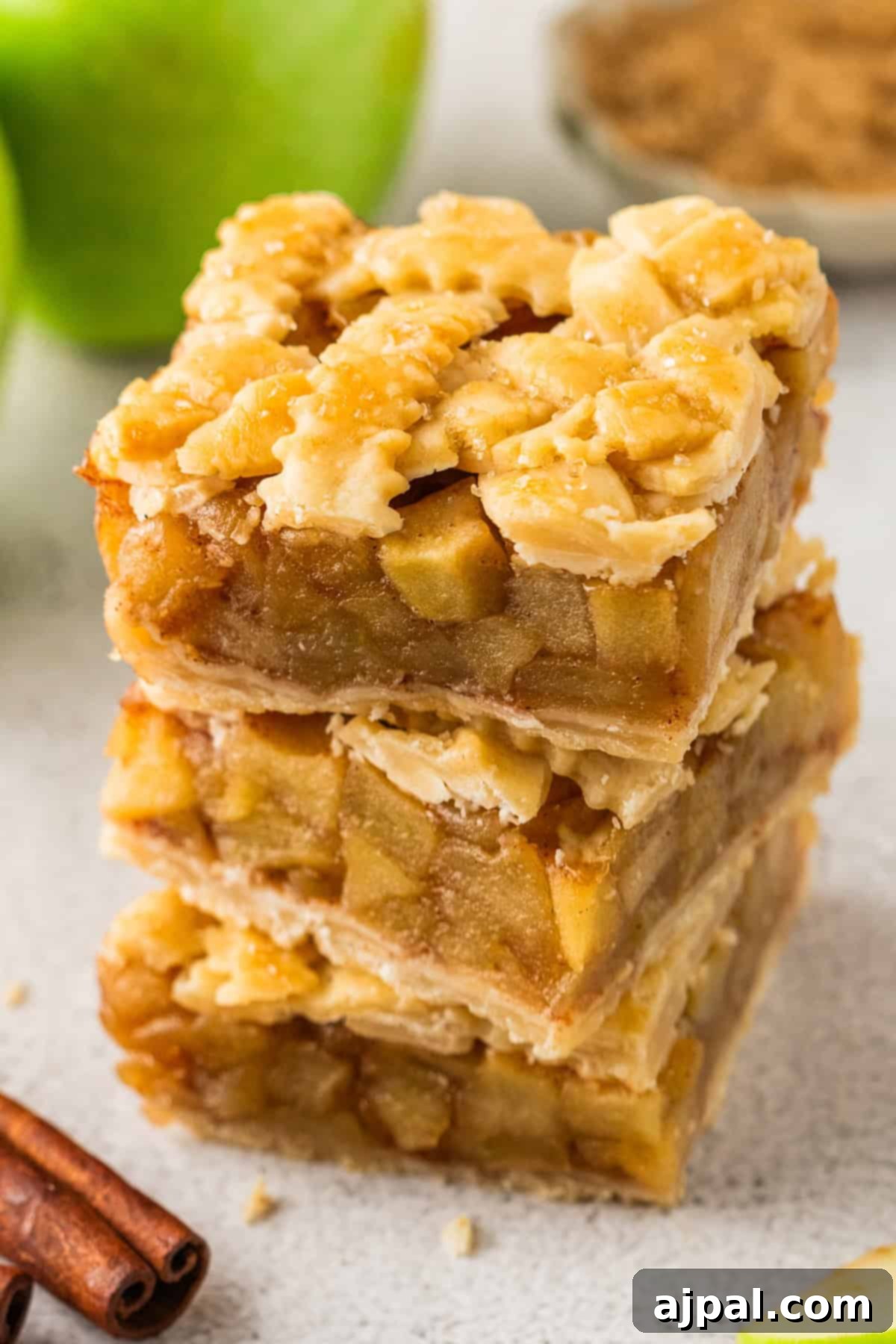 A stack of three Apple Pie Bars with a lattice crust, showing layers of diced apple filling and flaky pastry. Cinnamon sticks and a green apple are visible in the background.
