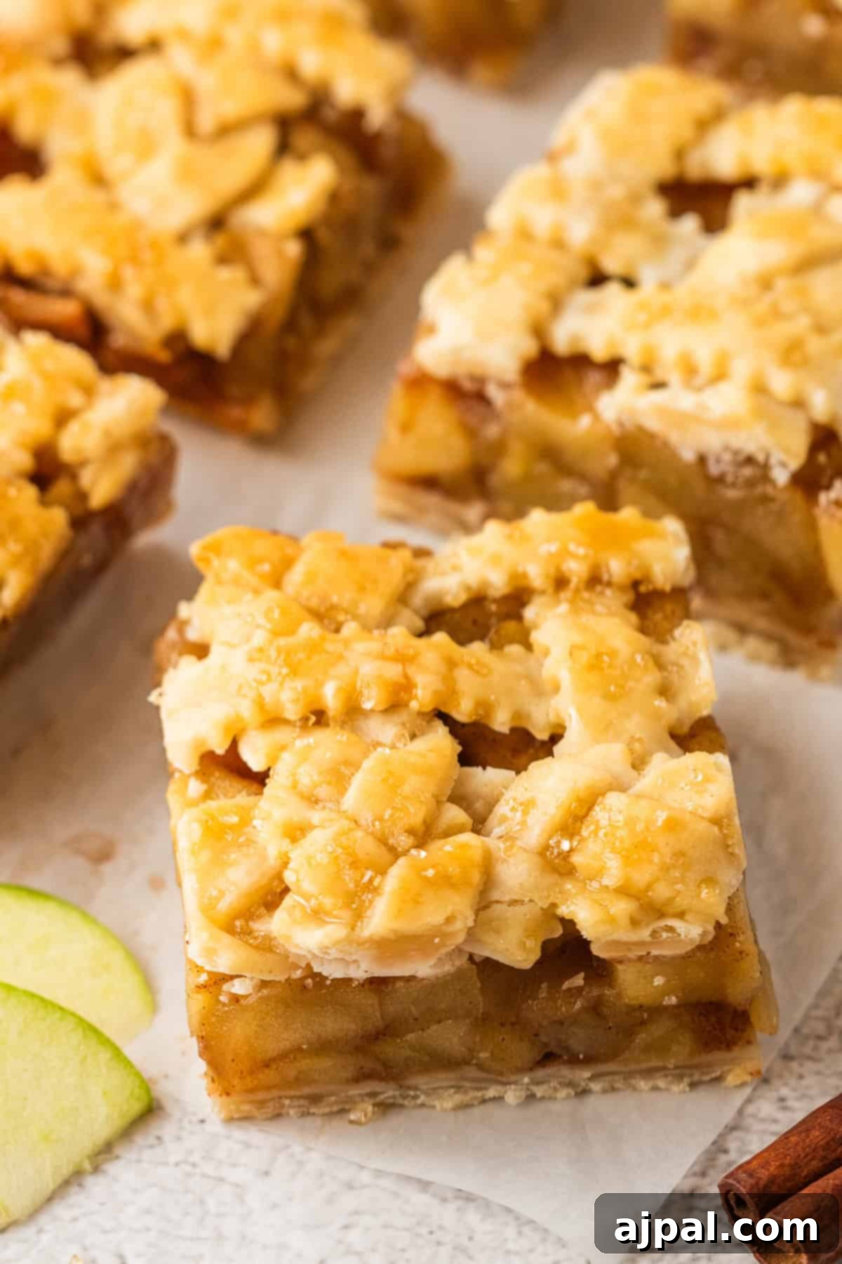 A close-up of Apple Pie Bars with a lattice crust, showing pieces of apple filling and a golden-brown, flaky topping. Slices of green apple are visible beside the bar.