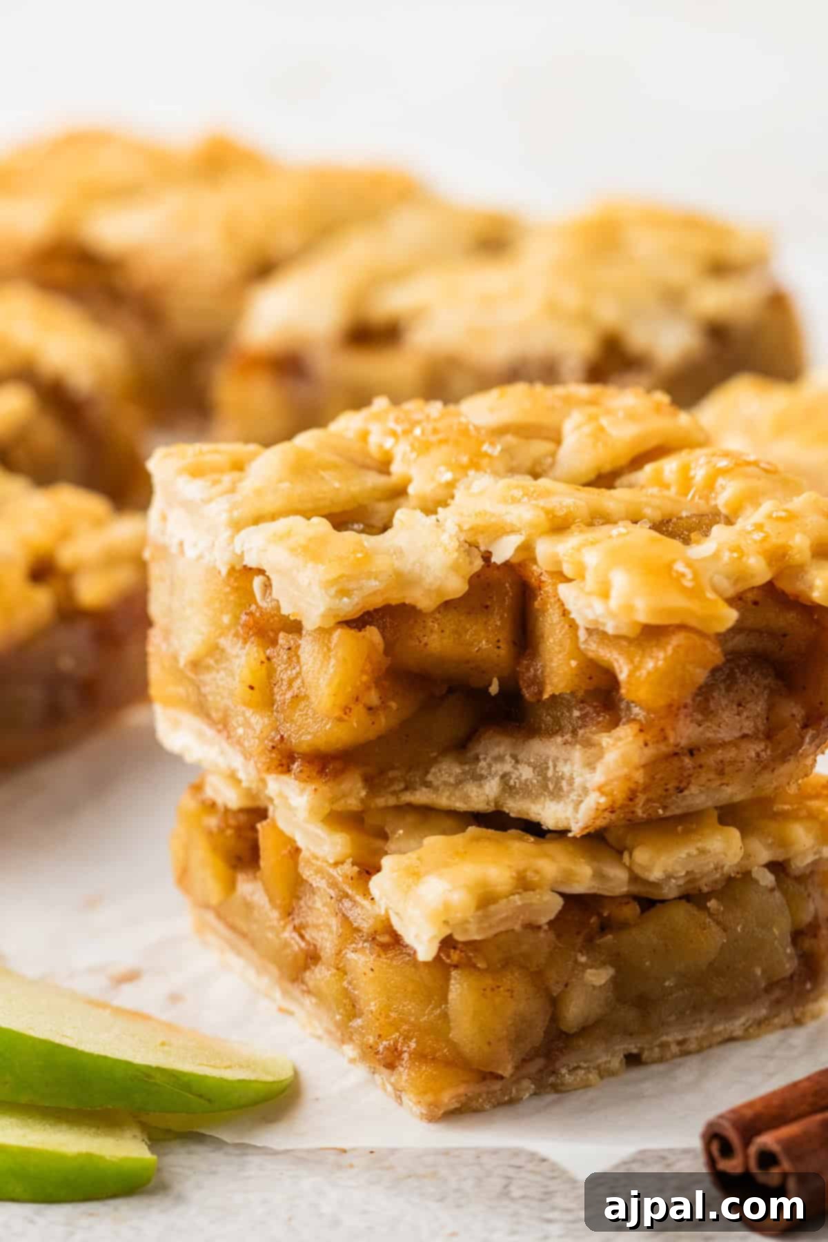 Two stacked Apple Pie Bars with a flaky lattice crust, filled with diced apples, sit on parchment paper. Slices of green apple and a cinnamon stick are visible nearby.