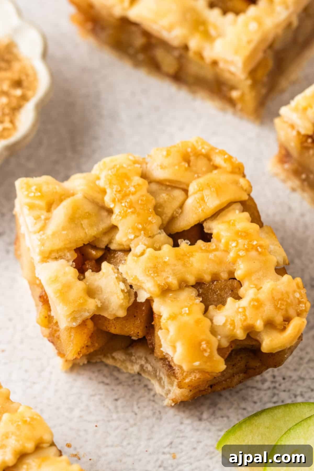 An Apple Pie Bar with a lattice crust and sugar topping sits on a light-colored surface, its square shape showcasing classic dessert charm.