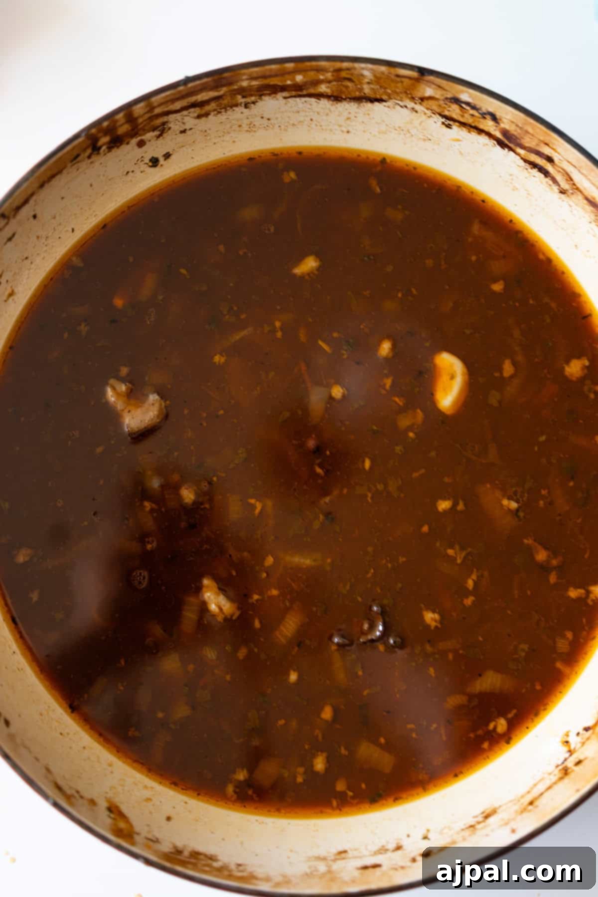A pot filled with dark brown broth, containing pieces of meat, herbs, and visible chunks of garlic.