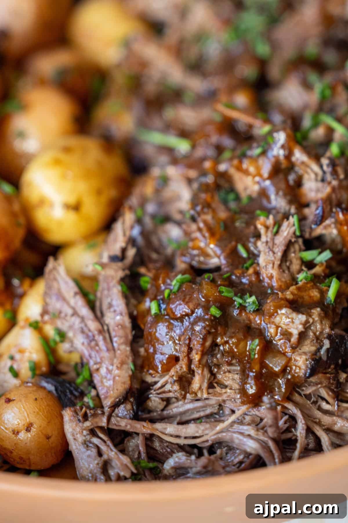 Close-up of shredded beef with brown sauce, garnished with chopped herbs, next to cooked baby potatoes.