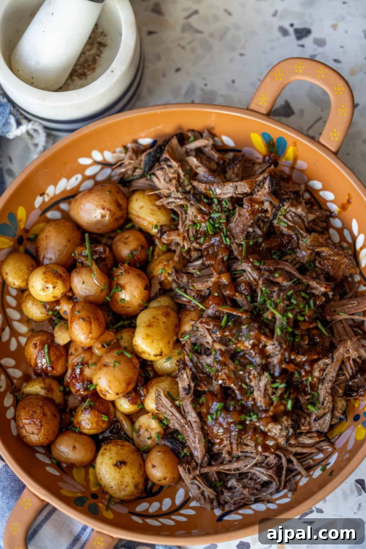 A ceramic dish with roasted baby potatoes and shredded braised beef garnished with chives, next to a mortar and pestle containing coarse salt.
