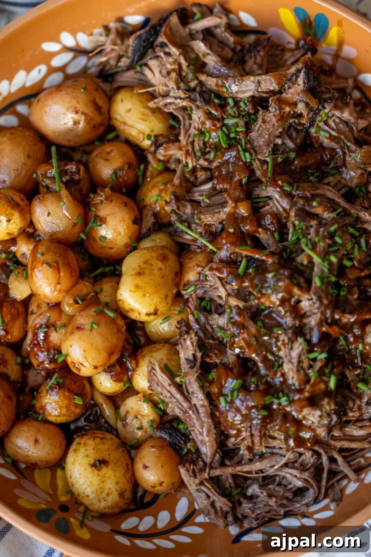 A dish of cooked baby potatoes and shredded beef, garnished with chopped herbs and drizzled with sauce, served in a brown patterned bowl.