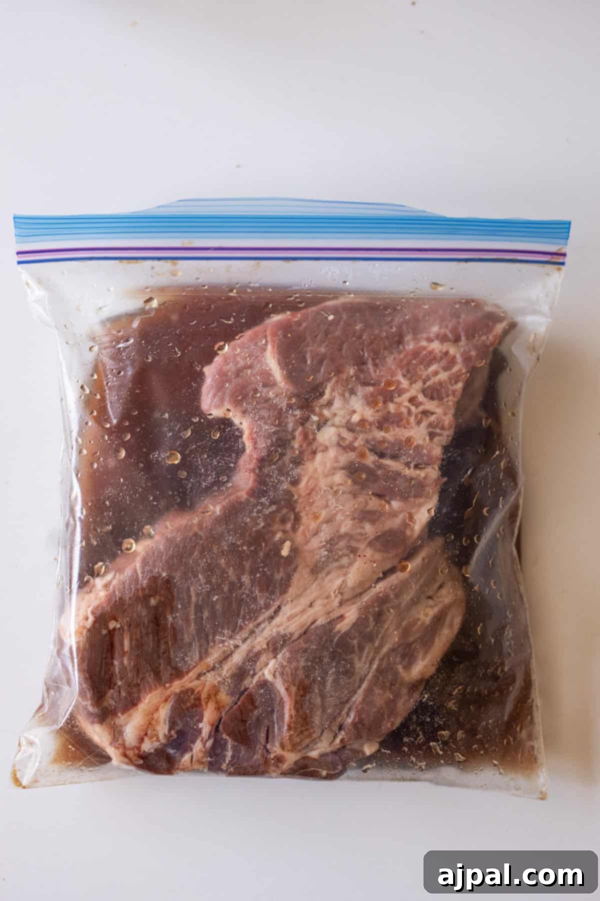 A large piece of raw meat marinating in a sealed plastic zip-top bag with a dark liquid, placed on a white surface.