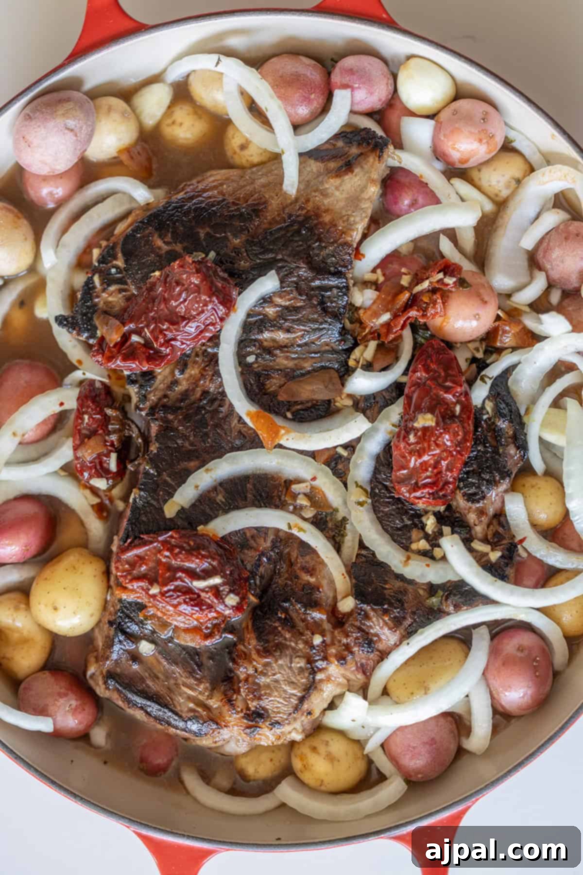 A pot with seared beef, red and yellow baby potatoes, sliced onions, whole garlic cloves, and sun-dried tomatoes, ready to be cooked.