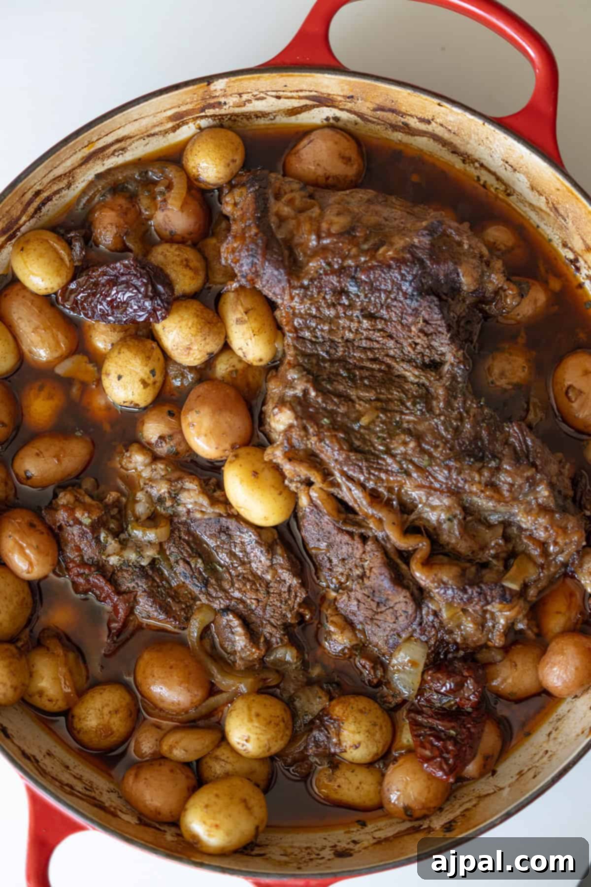 A red pot filled with cooked pot roast, baby potatoes, onions, and sun-dried tomatoes in a brown broth.