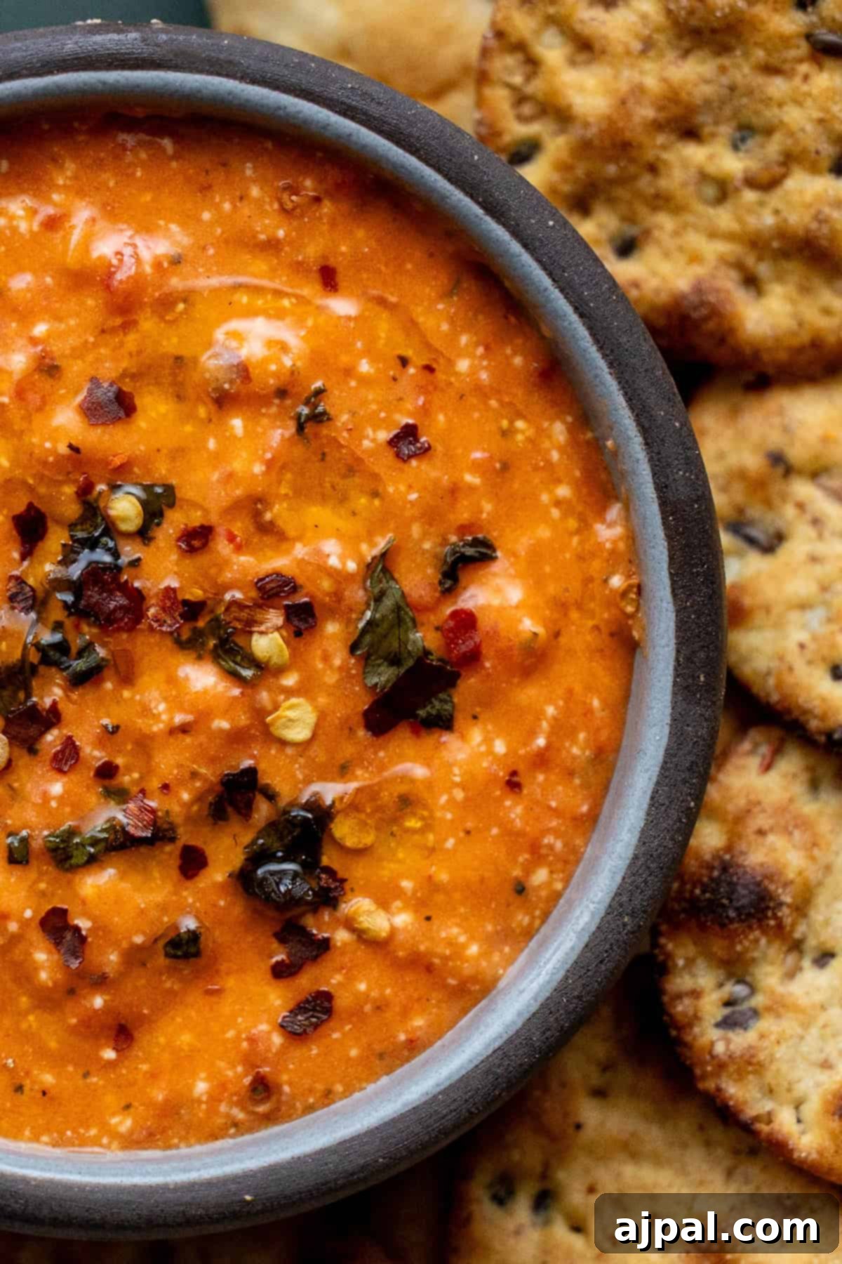 A bowl of creamy red dip garnished with herbs and chili flakes, surrounded by round, crispy crackers.