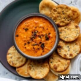 A bowl of red pepper dip garnished with chili flakes, surrounded by round crackers and mini flatbreads on a dark plate.