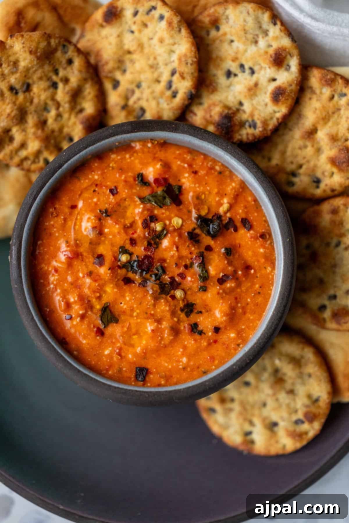 Red Pepper Feta White Bean Sensation 3 Close-up of a creamy, spicy red dip in a dark bowl, topped with a generous sprinkle of herbs and red pepper flakes, accompanied by artisan crackers.
