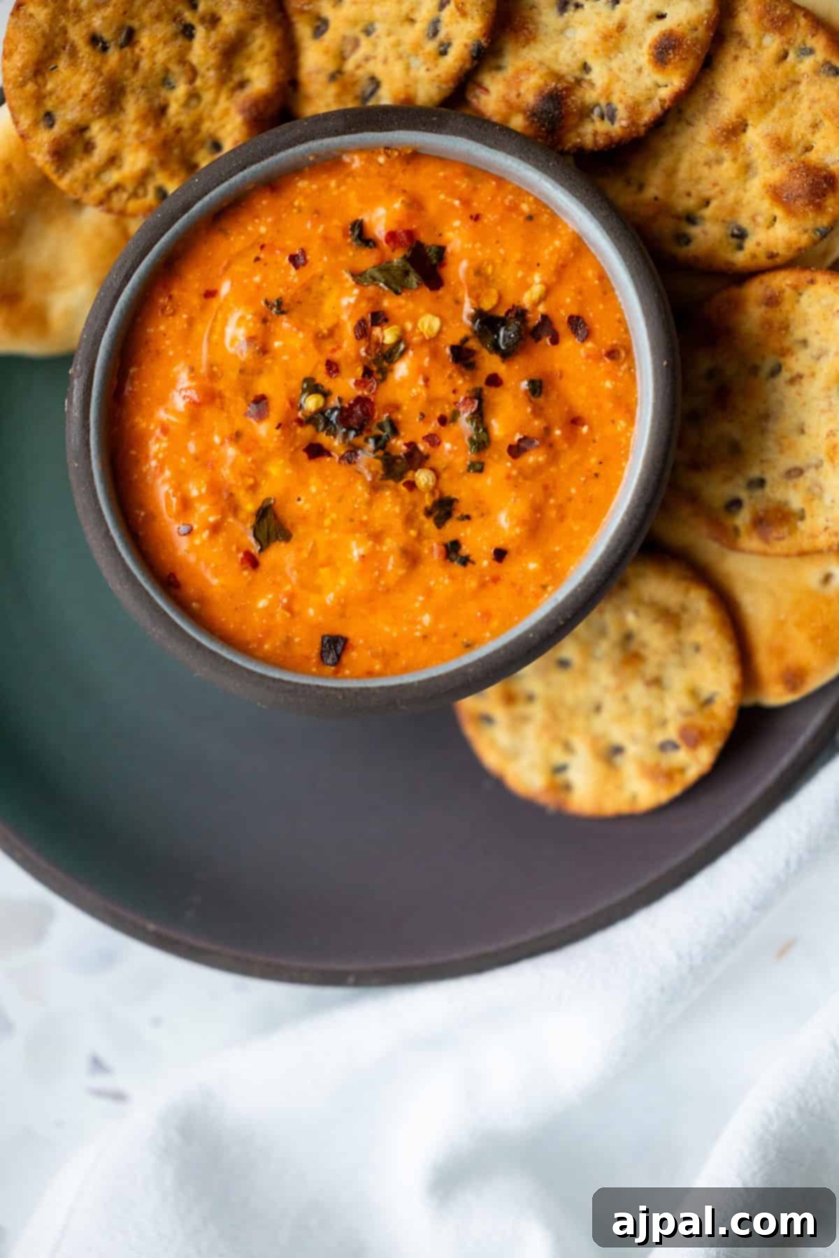 A bowl of red pepper dip garnished with chili flakes, surrounded by round crackers on a dark plate.