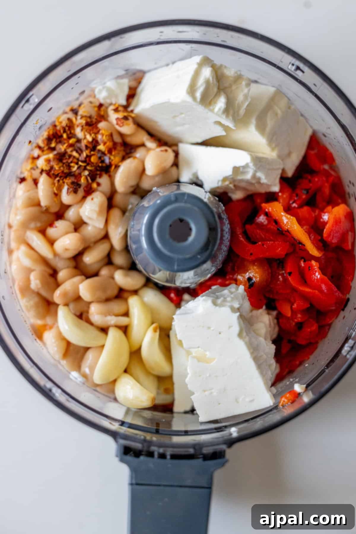 Food processor bowl containing white beans, garlic cloves, feta cheese, roasted red peppers, and red pepper flakes, viewed from above before mixing.