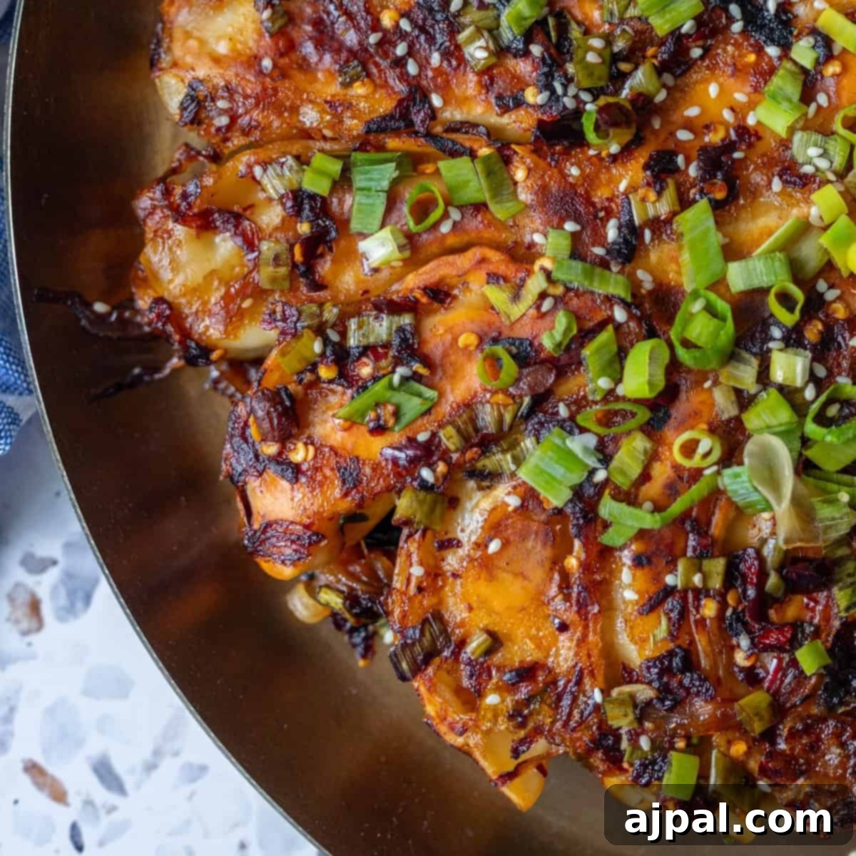 Fiery Potsticker Crispy Bake 11 Close-up of a savory dish featuring sliced potatoes topped with chopped green onions, sesame seeds, and a spicy, caramelized sauce on a gold plate—served alongside crispy potsticker pancakes.