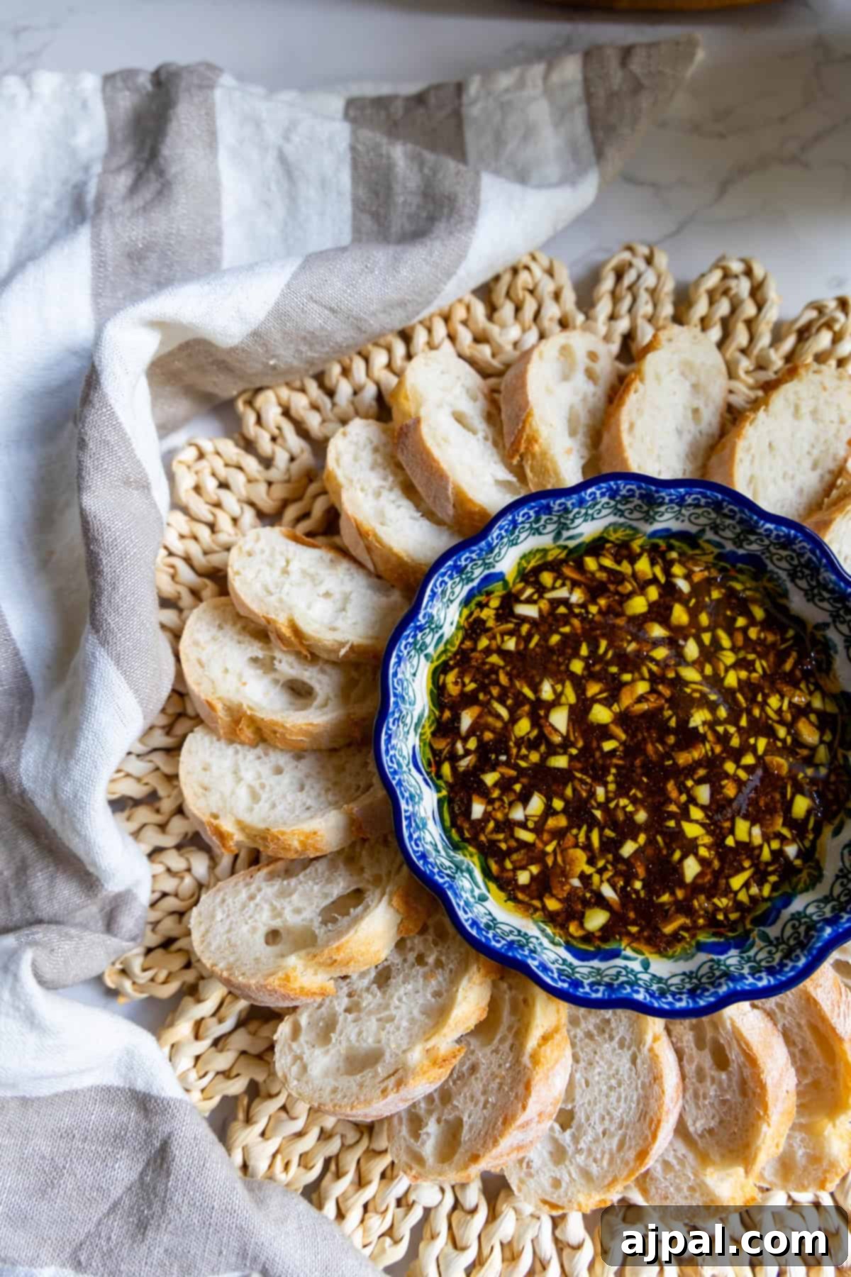 A bowl of chopped garlic and herb oil dip, reminiscent of a rustic balsamic vinaigrette, is surrounded by slices of baguette on a woven placemat with a striped cloth nearby.